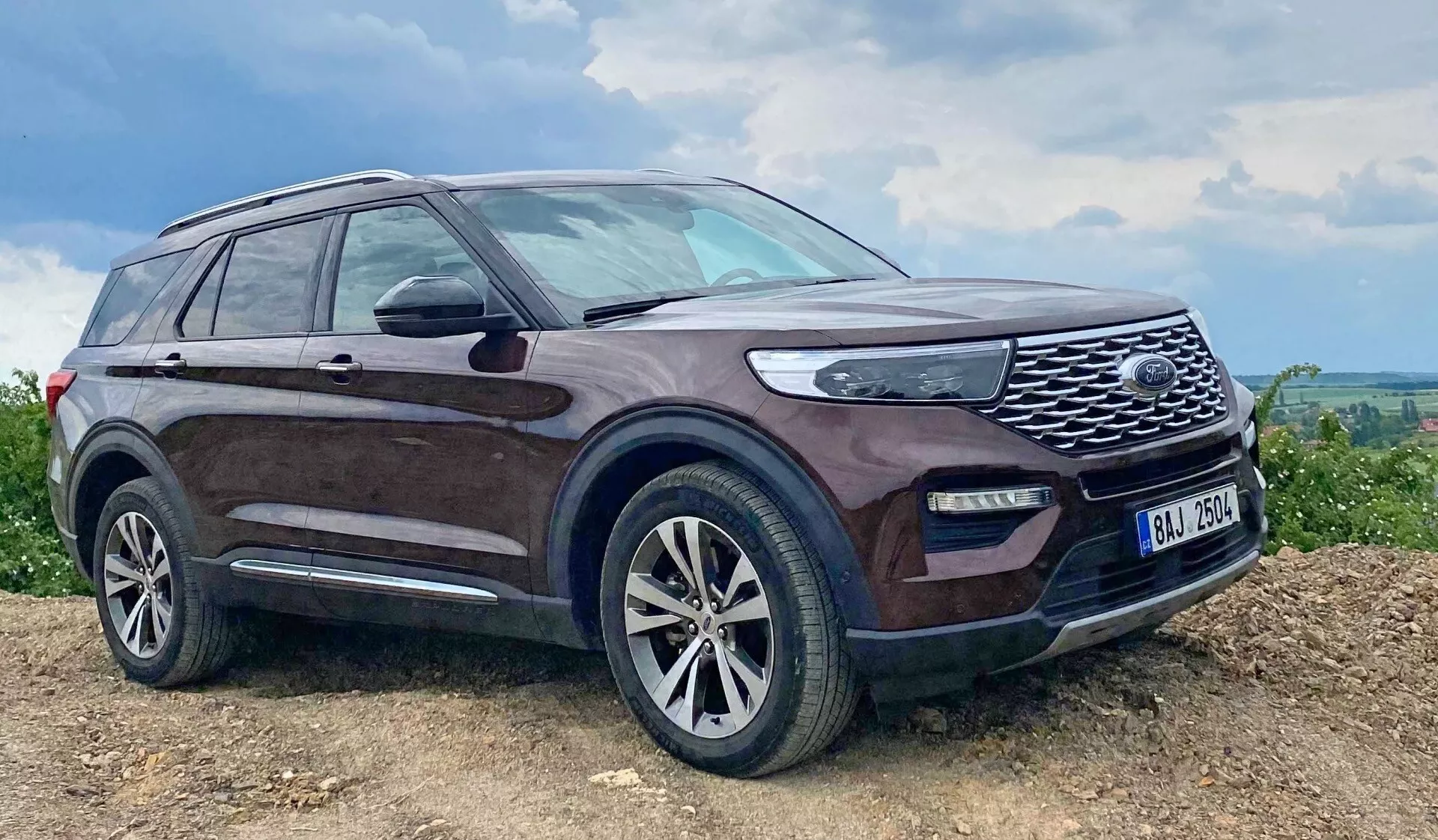 Image of a 2019 Ford Explorer SUV parked on an inclined, possibly unpaved surface featuring a dark hue with dirt spattered sides, prominent front grille with mesh design, LED headlights, fog lights, alloy wheels with multi-spoke design, side mirrors with integrated turn signals, roof rails, side step, slightly tinted windows, and an antenna on the rear roof, highlighting its rugged capabilities and luxurious features in a rural or off-road setting.