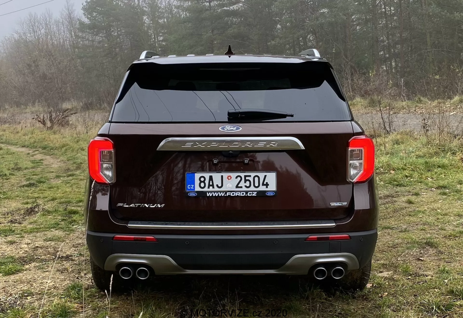 Rear view of a dark-colored 2019 Ford Explorer parked on a dirt path in a rural setting with vegetation and mist. The image showcases the clear-lens taillights, centrally placed Ford logo, 'EXPLORER' lettering, rear windshield wiper, parking sensors, dual exhaust system, and a trailer hitch cover. A license plate with a Czech URL is visible, emphasizing the vehicle's exterior design and technology.