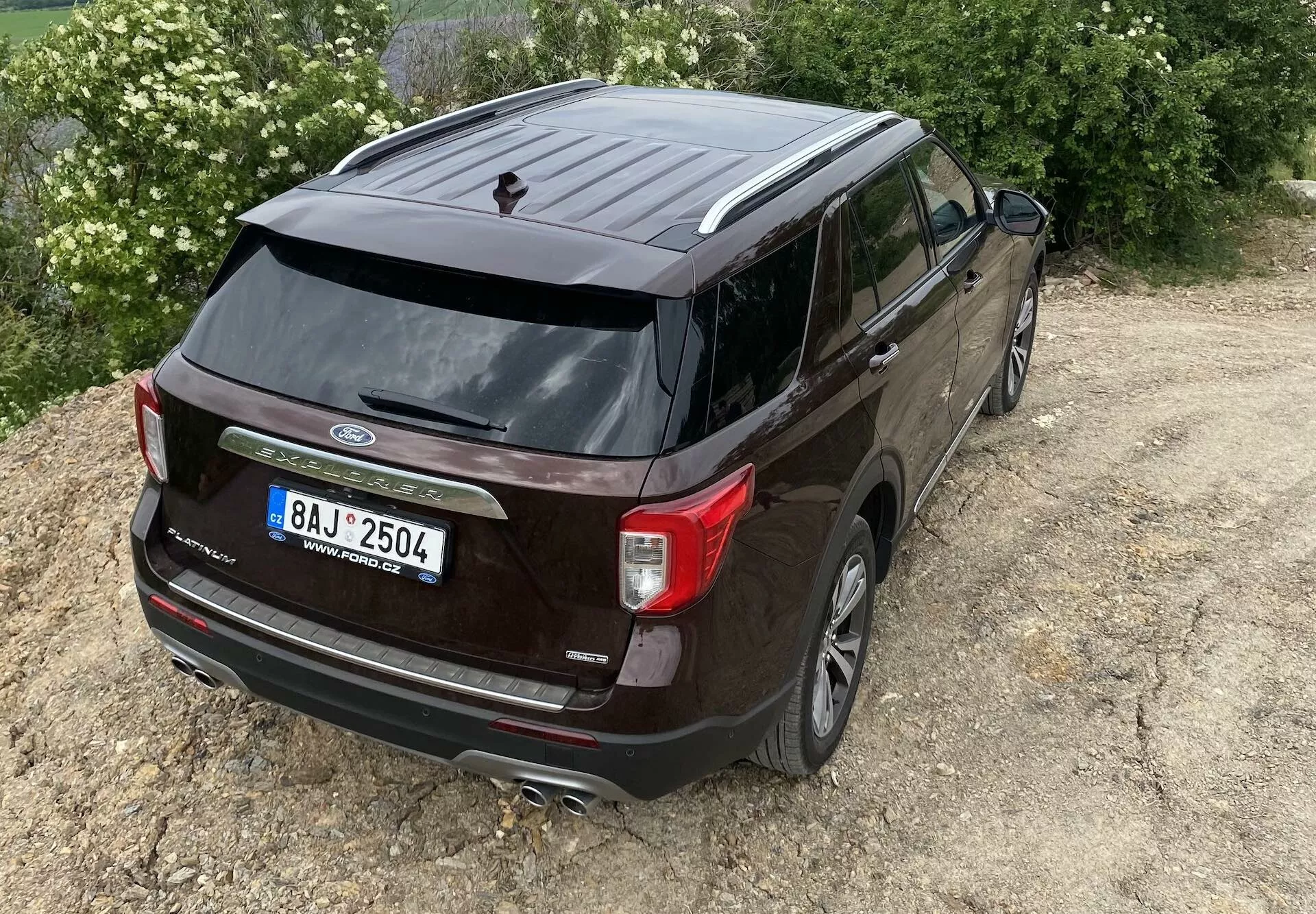 Elevated rear three-quarters angle view of a dark-colored 2019 Ford Explorer, highlighting the vehicle's rear end and side profile. Features visible include the Ford logo, model name 'Explorer', unique taillights, rear windshield wiper, shark-fin antenna, dual exhaust system, backup camera, alloy wheels, tinted windows, and side mirrors with integrated turn signal lights. The SUV is positioned off-center to the right on unpaved terrain, suggesting light off-road capability.