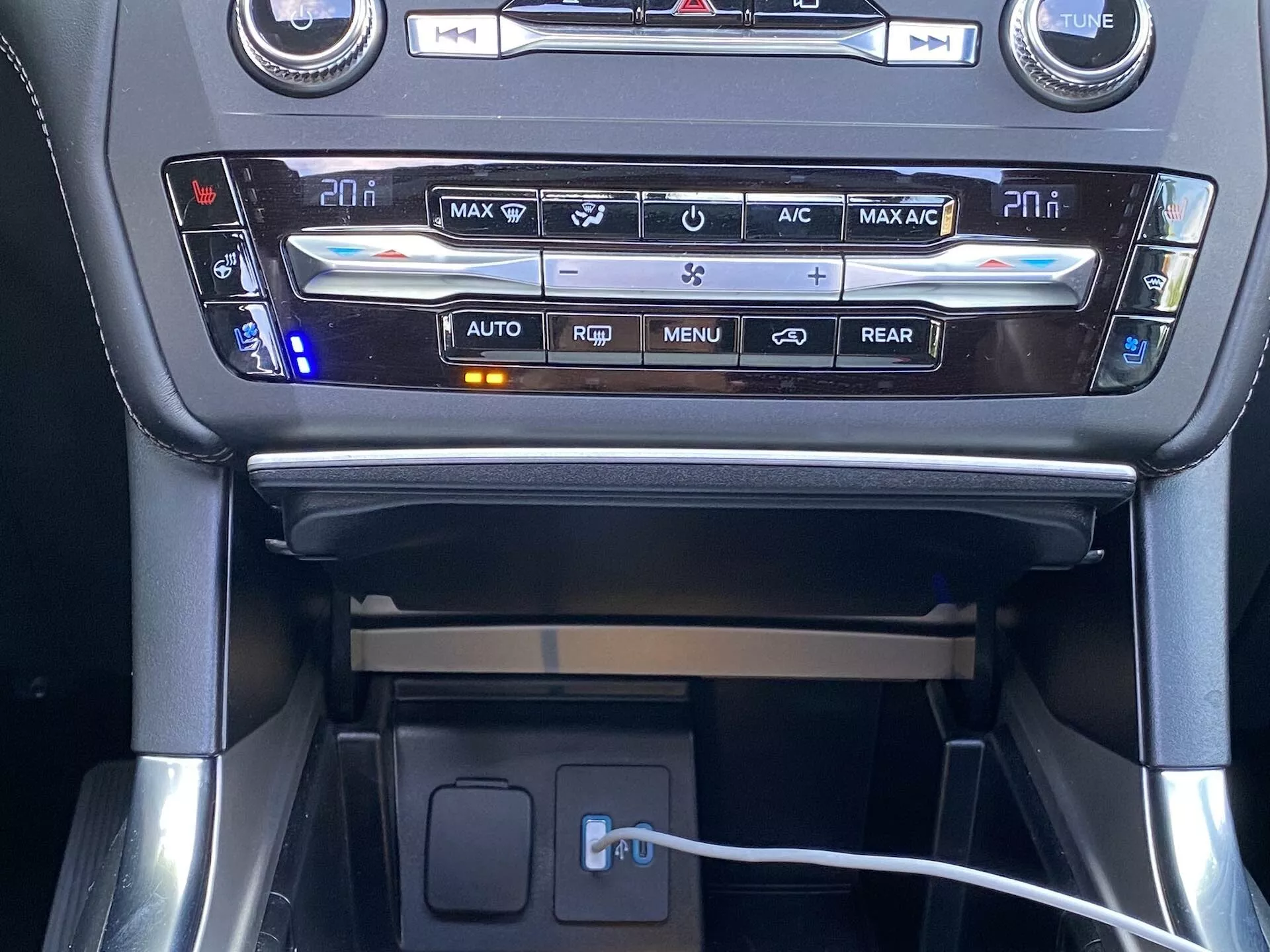 Frontal interior view of a 2019 Ford Explorer's center console, focusing on the climate control system and storage compartment. The well-lit climate control panel features digital temperature displays for driver and passenger, surrounded by buttons for various functions including defrost, air circulation, and automatic climate control. Below this panel, a small rubberized shelf with a visible charging cable indicates connectivity or charging features.