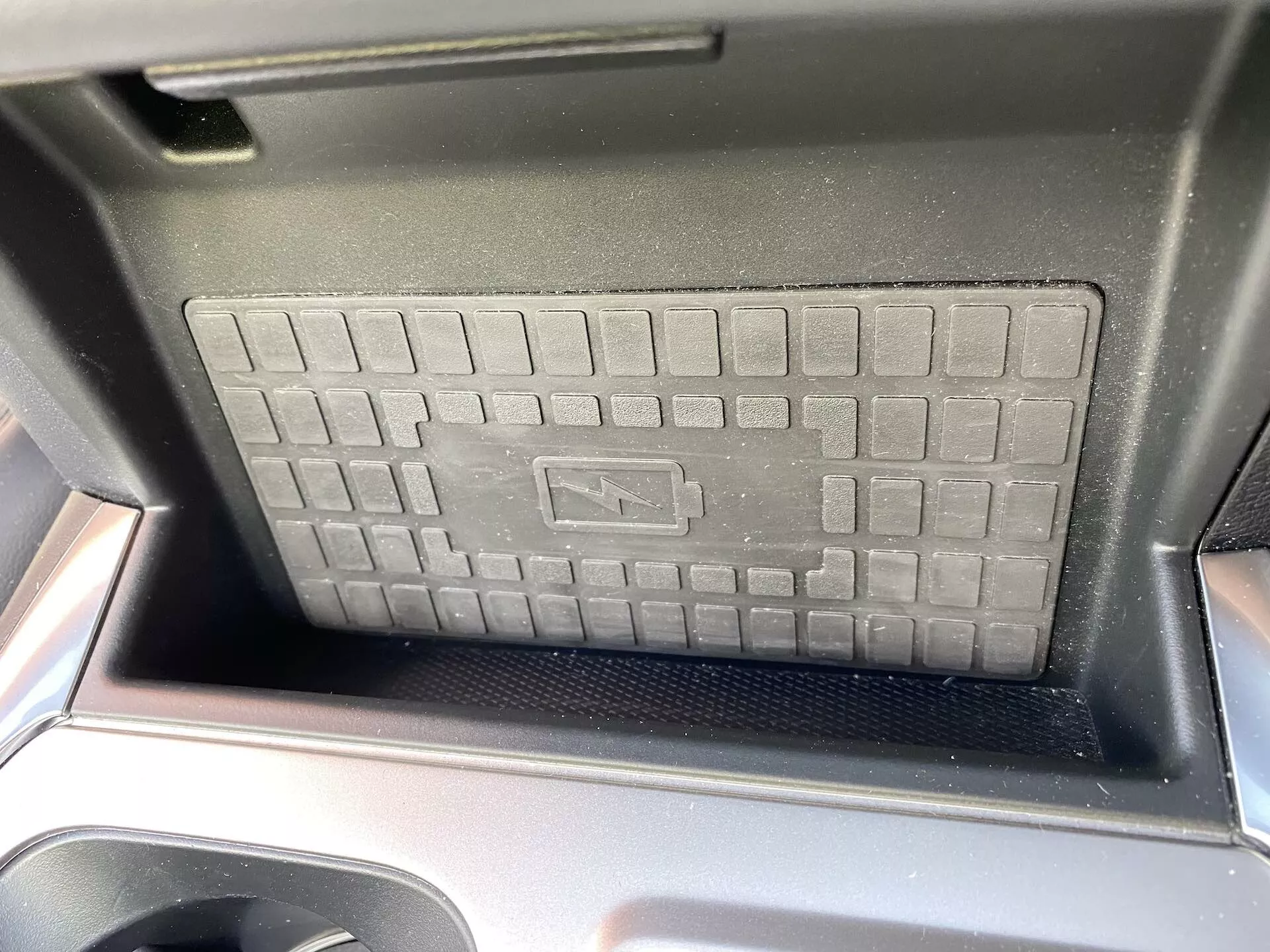 Close-up view of the Ford Explorer 2019's central console compartment, highlighting the wireless charging pad with a raised, grid-like texture and non-slip surface mat. The glossy black plastic surrounds are visible, adding a touch of modern automotive design. Sunlight suggests daytime photo from the front seat, with the charging pad symbol denoting advanced wireless charging technology.