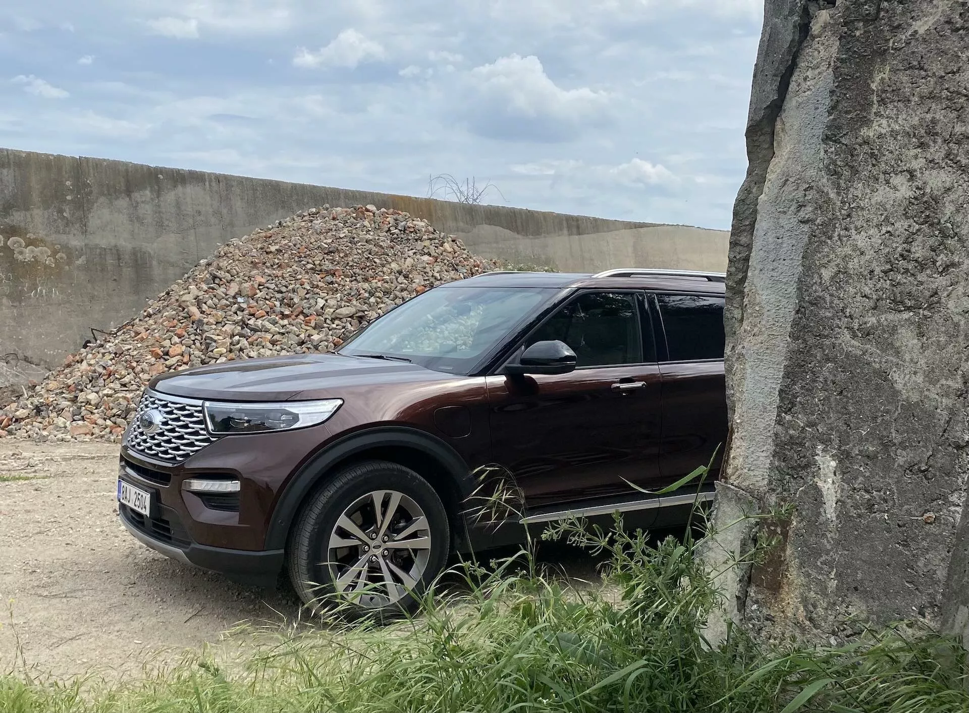 The image showcases a 2019 Ford Explorer SUV from a three-quarter front view, highlighting the front left grille, sleek LED headlights, hood, front wheel, and fender. The vehicle, with a glossy deep maroon or burgundy color, stands in an outdoor setting with gravel in the background, suggesting a construction site. Alloy multi-spoke wheels, integrated turn signal wing mirrors, and chrome accents add to its premium look. Partially obscured by a concrete structure on the right, this SUV features modern design and safety elements, making it suitable for categories like SUVs, modern car design, vehicular technology, and off-road vehicles.