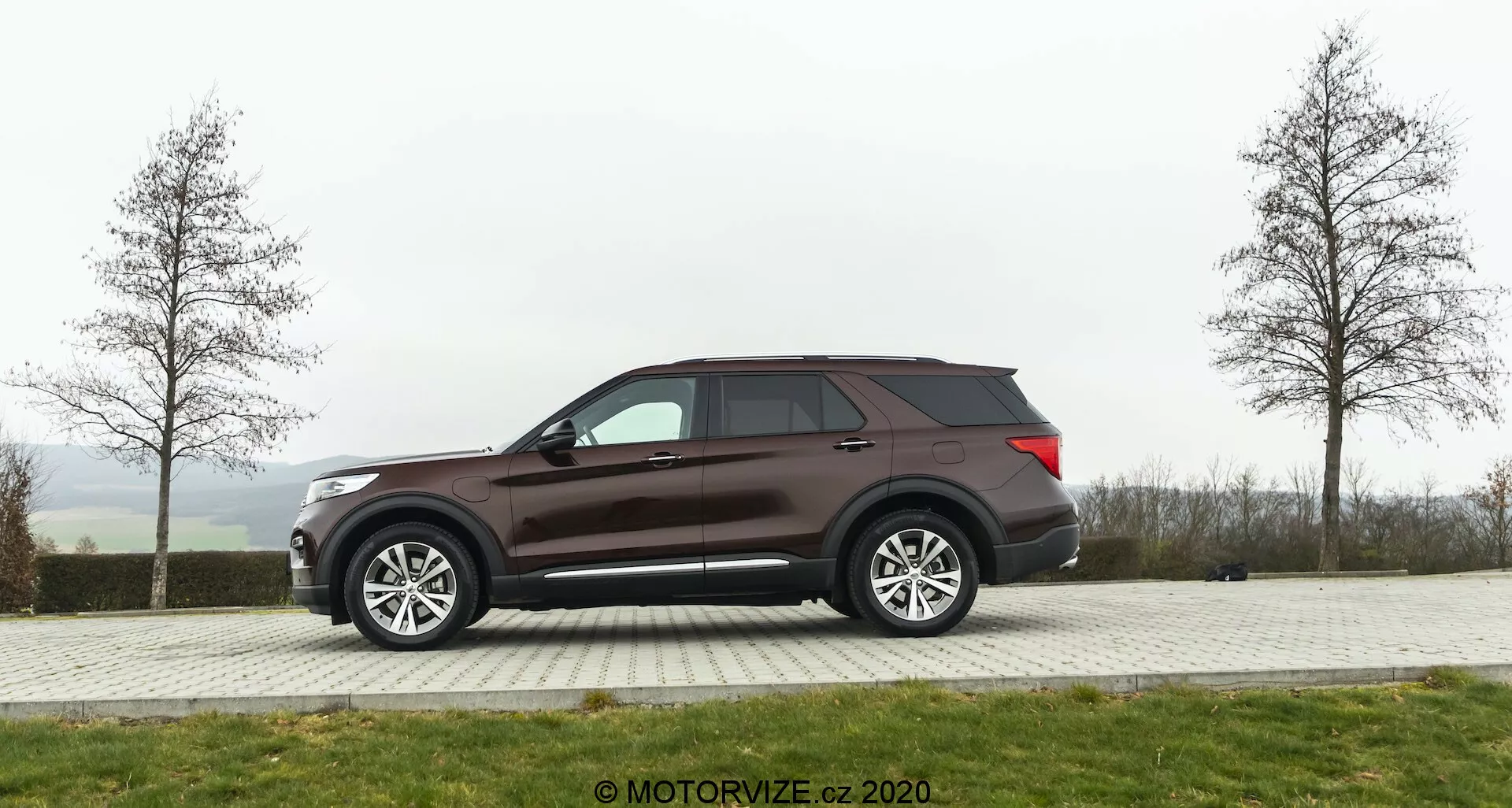 Side profile view of a 2019 Ford Explorer SUV parked on a paved area with grass in the background on an overcast day. The vehicle features large multi-spoke alloy wheels, tinted windows, and a dark brown or burgundy paint job. Design elements include side mirrors likely with power adjustment and turn signal integration, aerodynamic roofline, and a visible rear antenna.