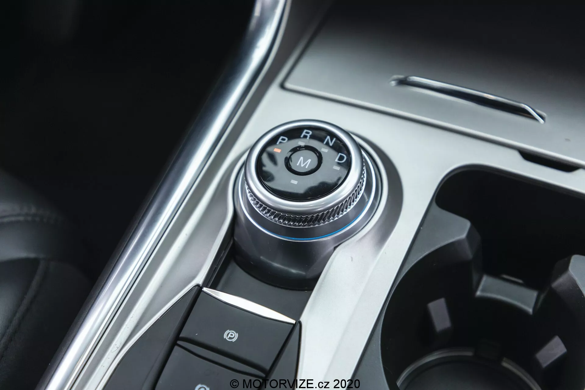 Close-up view of the gear selector knob and part of the center console of a 2019 Ford Explorer. The focal point is the rotary dial style shifter with 'P, R, N, D' labels indicating the automatic transmission system. The photo is taken from a slightly off-center, top-down angle. Surrounding the gear selector are buttons, including one with a parking brake symbol, suggesting an electronic parking brake system. The console features a combination of metallic and plastic finishes, emphasizing the premium interior of the vehicle. The well-lit image highlights the metallic elements, with a shallow depth of field focusing on the gear selector.