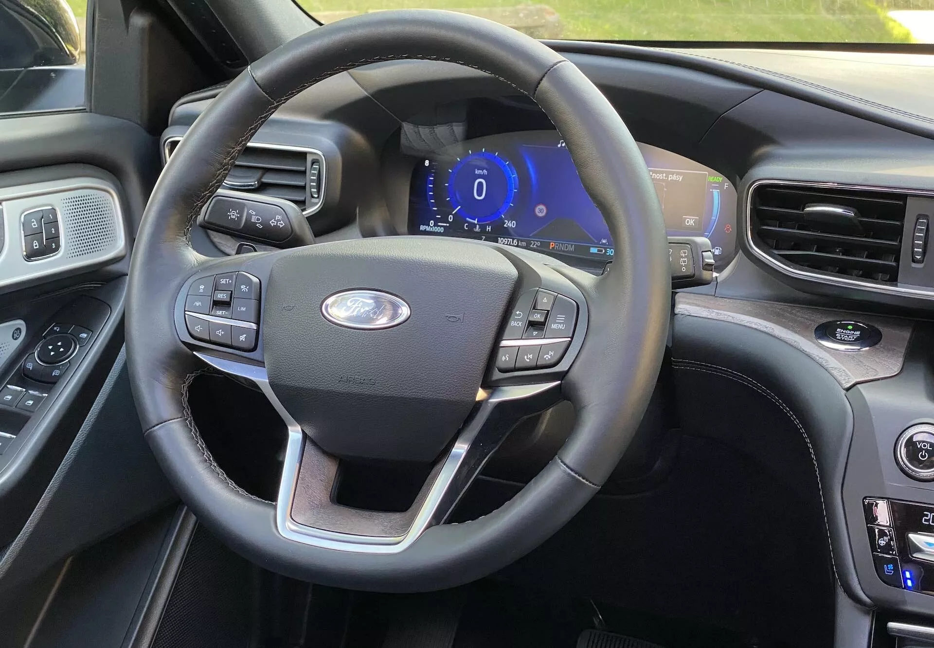 Interior view of a 2019 Ford Explorer from the driver's seat, showcasing a three-spoke steering wheel with multifunctional buttons for cruise control, audio settings, and phone connectivity. The centrally located Ford emblem and a blue-illuminated digital instrument panel suggest the vehicle is in accessory or start-up mode. Visible to the right is part of the center console with climate control buttons and temperature settings, indicating dual climate control. The interior features a mix of leather and wood grain finishes for a premium feel, with another larger display screen slightly visible at the edge.