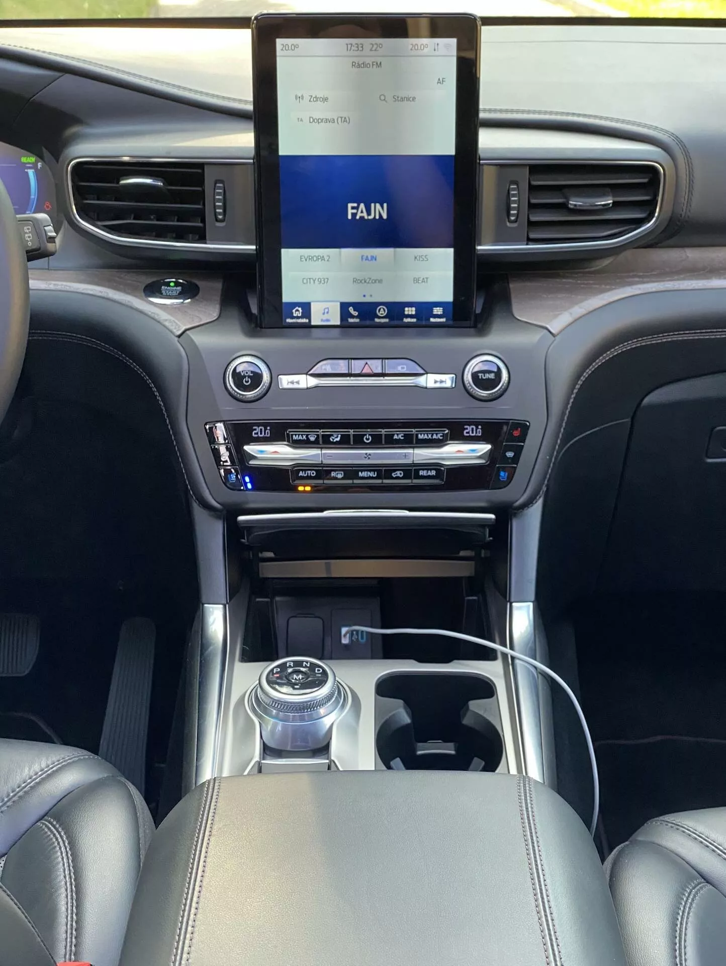 Interior view of a 2019 Ford Explorer from the perspective of a rear seat passenger, focusing on the front cabin area with an advanced infotainment system on the center console, dual-zone climate control, rotary gear shift knob, and luxurious leather and chrome trim details.