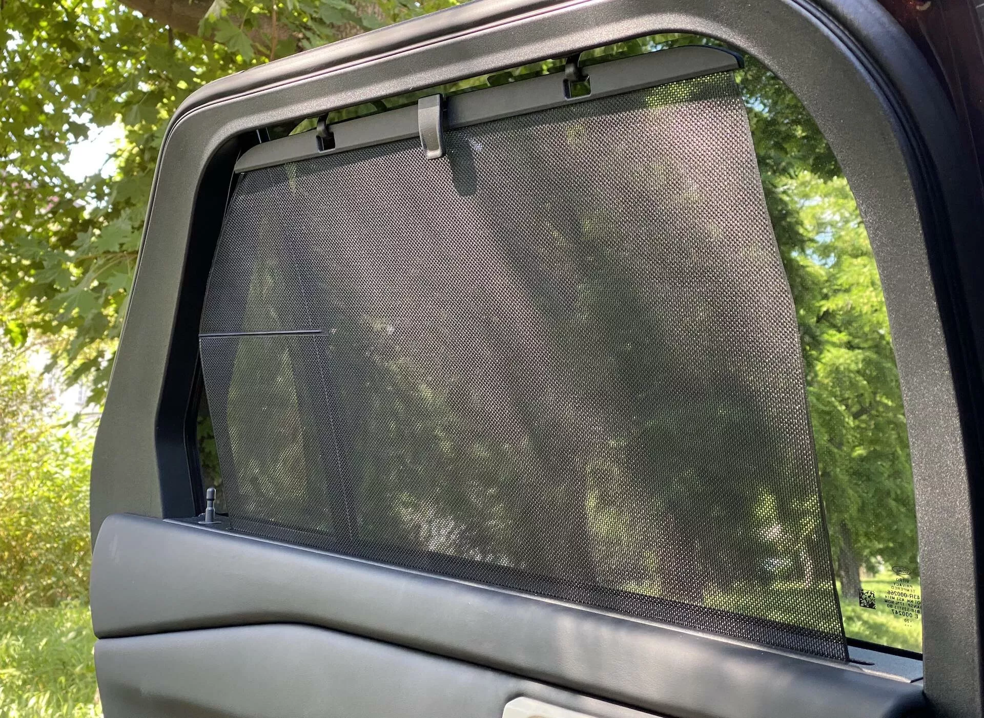 Close-up interior view of the 2019 Ford Explorer's rear side door and window. The image captures a partially lowered window with a mesh sunshade installed, covering the entire glass area and extending slightly beyond its top edge. The upper portion of the car door panel, featuring a smooth trim, is also visible. Taken from a horizontal orientation, the photo shows light filtering through the sunshade, suggesting a park-like outdoor setting. Focus on passenger comfort with sun protection accessory.
