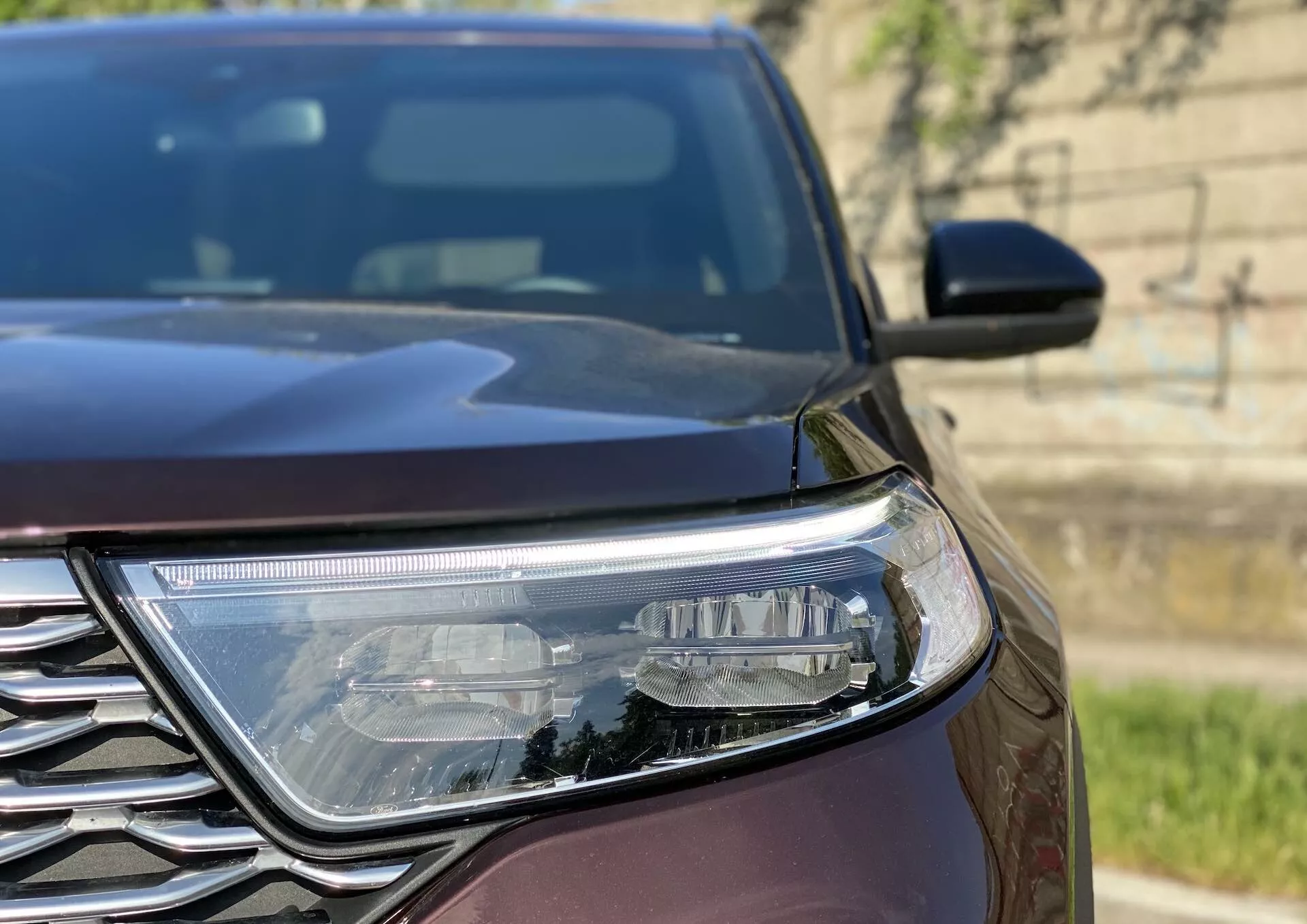 Close-up, angled view focusing on the left-side headlamp and portion of the grill of a 2019 Ford Explorer, highlighting the angular design lines of the LED headlamp and intricate chrome-trimmed grill pattern in natural daylight.