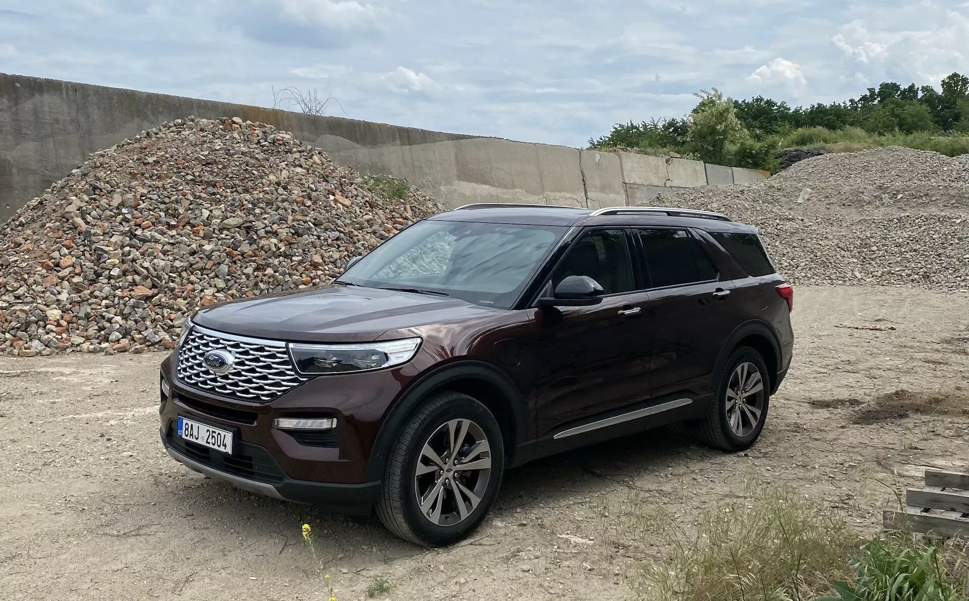 The image shows a 2019 Ford Explorer, a modern SUV, parked on uneven terrain resembling a construction site or off-road area. Captured from a three-quarter perspective, the photo showcases the car's front and side, including the grille, headlights, front bumper, hood, alloy wheels, front and rear doors, side mirrors with integrated turn signals, fender, and windows. The car is oriented facing right and the metallic paint finish is clearly visible, providing a comprehensive view of this well-maintained vehicle with roof rails, suggesting additional cargo capacity.