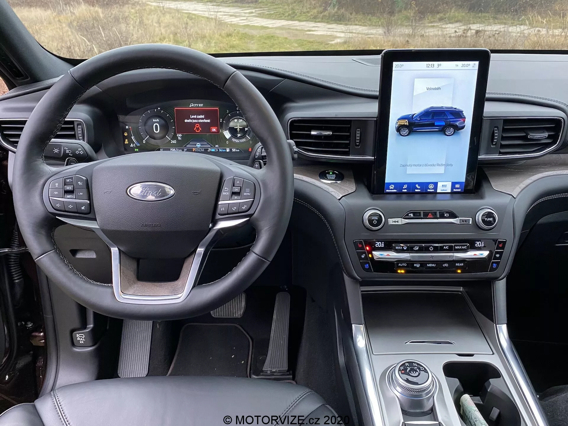 Interior of a Ford Explorer 2019 focusing on the front cabin from the driver’s seat perspective. The image showcases a steering wheel with control buttons for volume, voice commands, and cruise control, alongside a Ford emblem. The digital instrument panel displays a speedometer, tachometer, drive mode, and fuel status. The center console features a large vertical touchscreen for the infotainment system, displaying a blue vehicle illustration, traditional controls for climate and audio settings, and a gear shift knob. The dashboard has a contemporary design with glossy surfaces, matte finishes, and brushed metal accents, enhancing the modern, premium feel. Technological features include a multifunction steering wheel, digital instrument cluster, touch-enabled infotainment system, automatic climate control, drive mode selector, and seat temperature control.