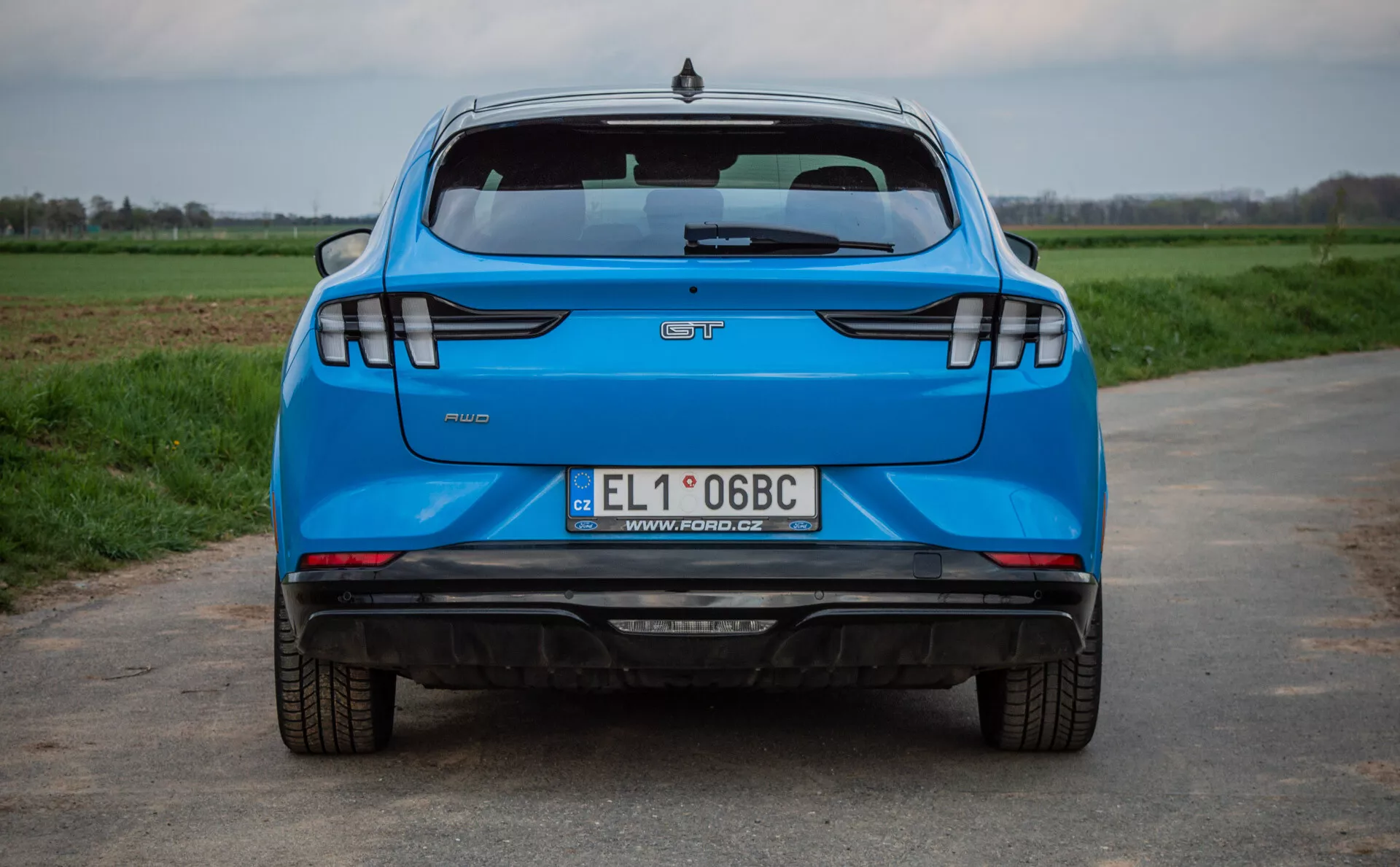 Rear view of a blue 2020 Ford Mustang-Mach-E on an asphalt road with grass fields in the background, showcasing key features like the manufacturer logo, model name, trim level badge, Polish registration license plate, double exhaust system, modern LED taillights, parking sensors, and a high-mounted stop lamp.