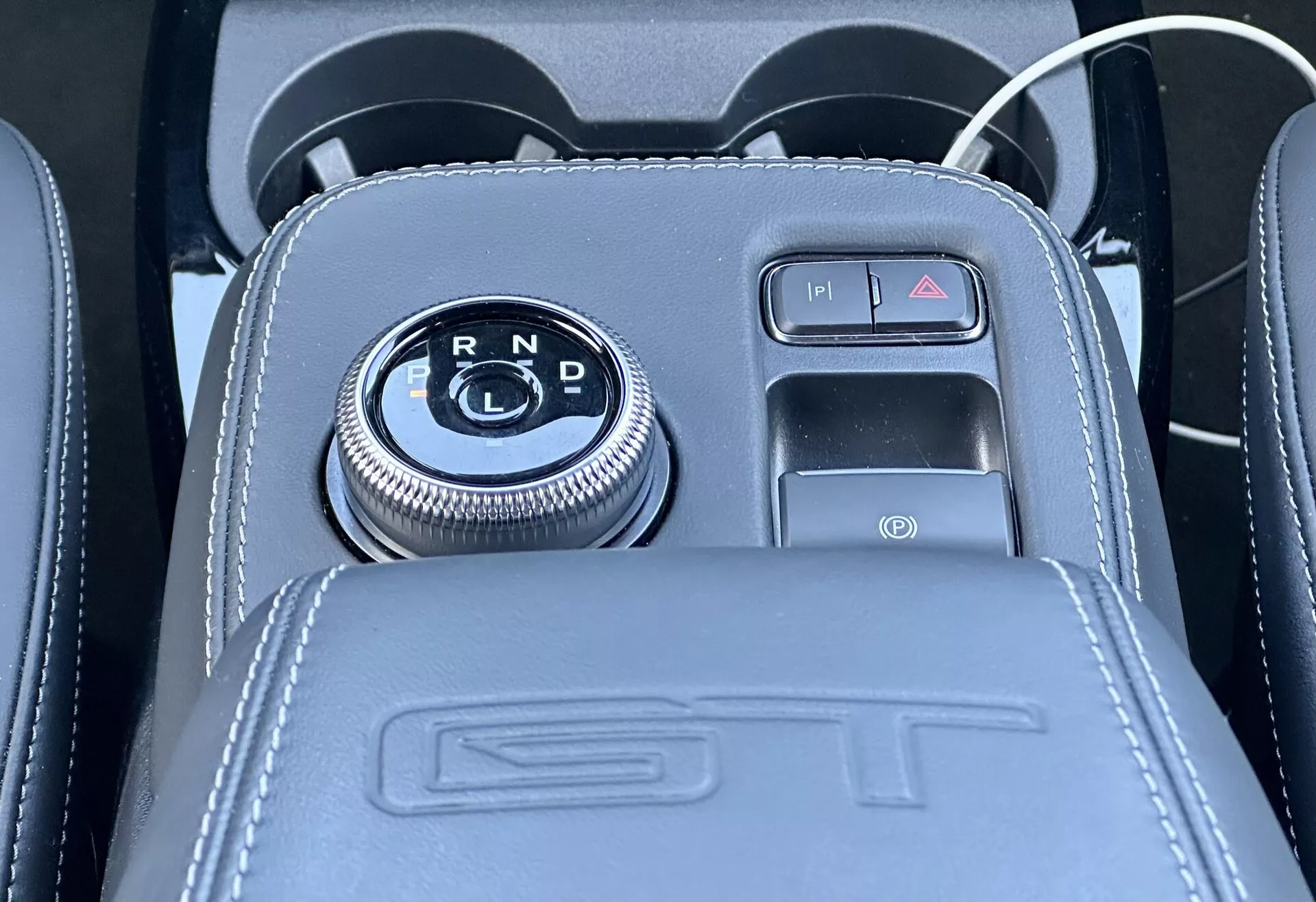 Close-up view of Ford Mustang Mach-E 2020 center console featuring a rotary gear selector set in a glossy black panel with R, N, D gear indicators, surrounded by a chrome finish, next to a push-button electronic parking brake with leather upholstery detailing, ST embossing, and including a USB port and 12V accessory socket