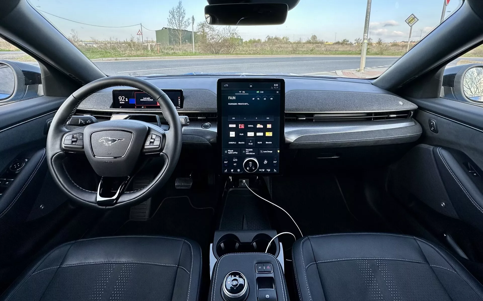 First-person view of Ford Mustang Mach-E 2020 interior, showcasing the left-hand position steering wheel with flat-bottom design and emblem, digital instrument cluster, large vertical touchscreen infotainment system on center console with climate control knobs below, USB ports, and luxurious dark-toned leatherette upholstery with detailed stitching, hinting at advanced safety and assistive features, with a rural exterior backdrop visible through the windshield.