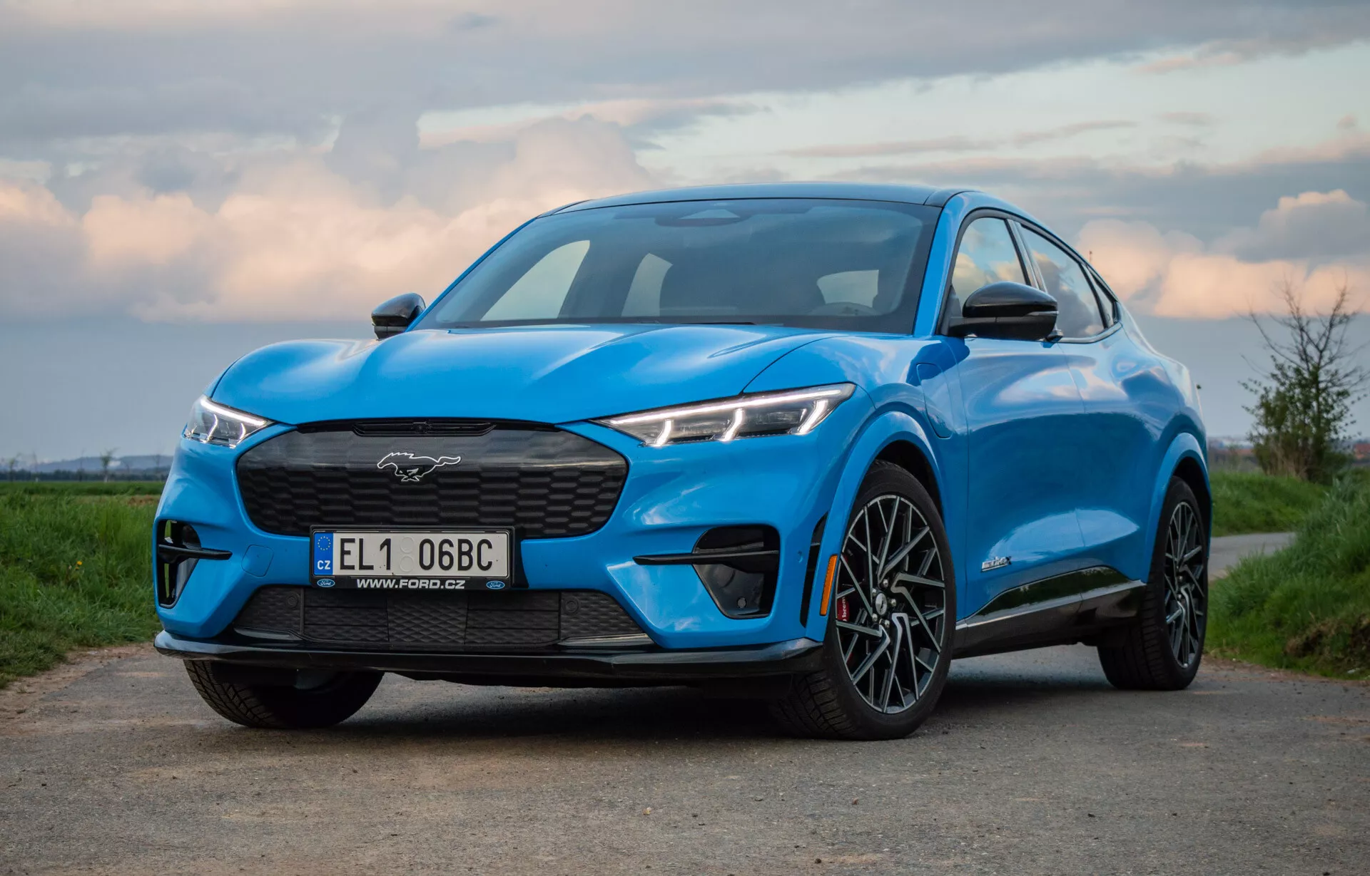 Blue 2020 Ford Mustang Mach-E photographed at low and slightly off-center angle showcasing the electric SUV's grille, headlamps, hood, and profile including front wheel and fender, emphasizing its performance and aerodynamic design at dawn or dusk.