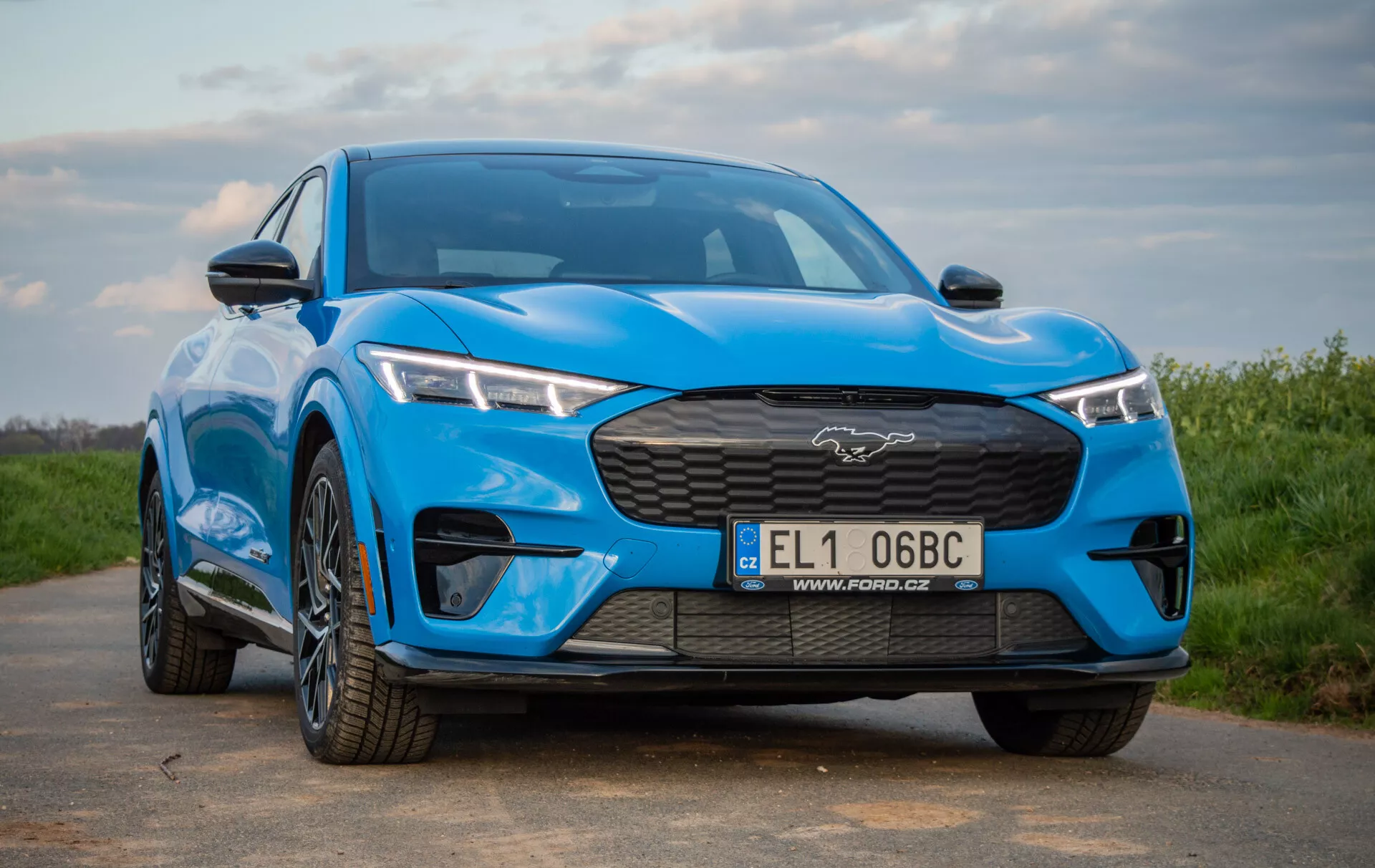 Close frontal perspective of a blue Ford Mustang Mach-E 2020 showcasing its distinctive closed grille with a large Mustang emblem, sleek LED headlights, and sporty front bumper, emphasizing the vehicle's all-electric crossover design and aerodynamic features.