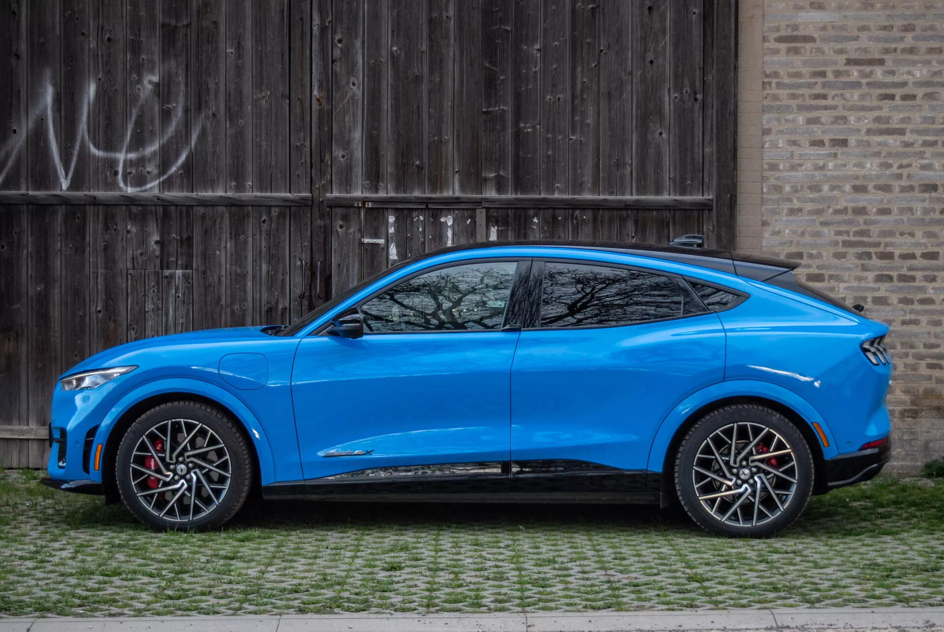 2020 Ford Mustang Mach-E blue crossover SUV parked parallel to a rustic wooden garage door with graffiti, profile view showcasing aerodynamic design, coupe-style sloping roofline, four-door model with integrated handles, aerodynamically styled side mirrors with turn signals, large two-tone alloy wheels with red brake calipers, moderate ground clearance, contoured body with sculpted lines and pronounced wheel arches, tapered window line with small quarter window after the C-pillar, and sporty side skirts