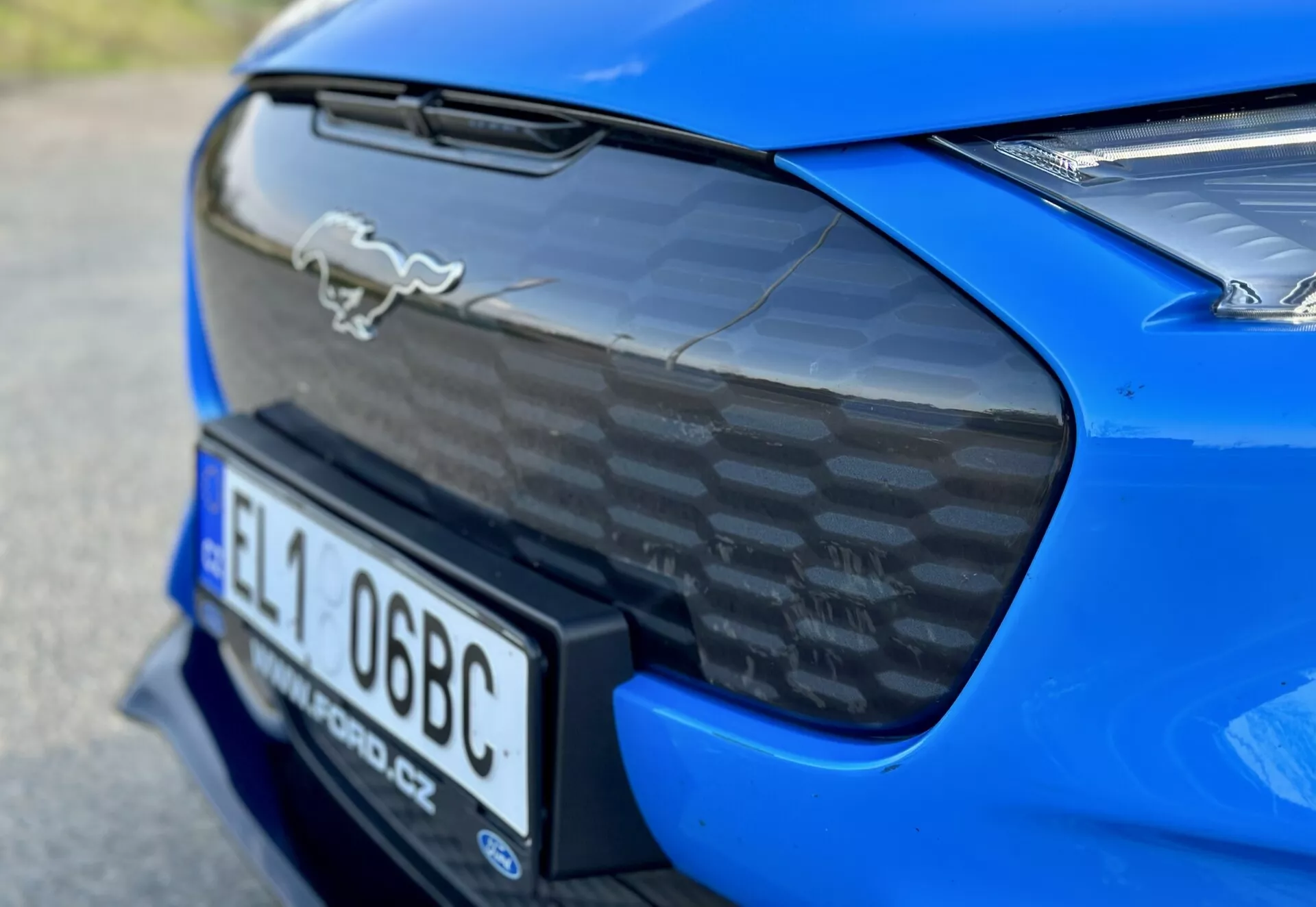 Close-up of the rear section of a 2020 Ford Mustang Mach-E in blue, showcasing the chrome horse emblem, carbon fiber rear decklid spoiler, integrated rear registration plate nook, and partial view of LED taillights, accentuating the car's aerodynamics and performance features.
