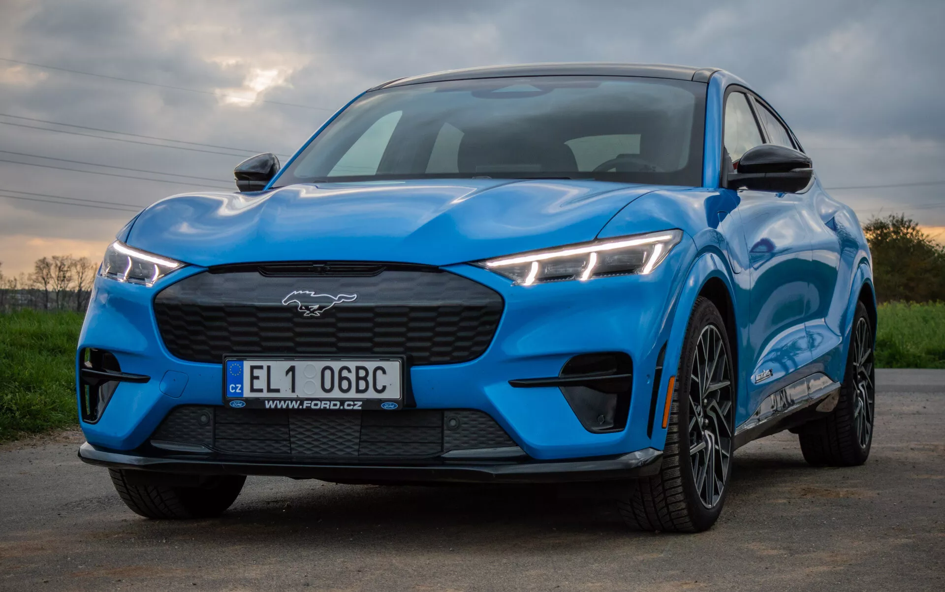 2020 Ford Mustang Mach-E in bright blue exhibiting a sporty and aerodynamic design from a low front three-quarter perspective, highlighting angular LED headlights, closed grille, contoured hood, and multi-spoke alloy wheels against a natural backdrop.