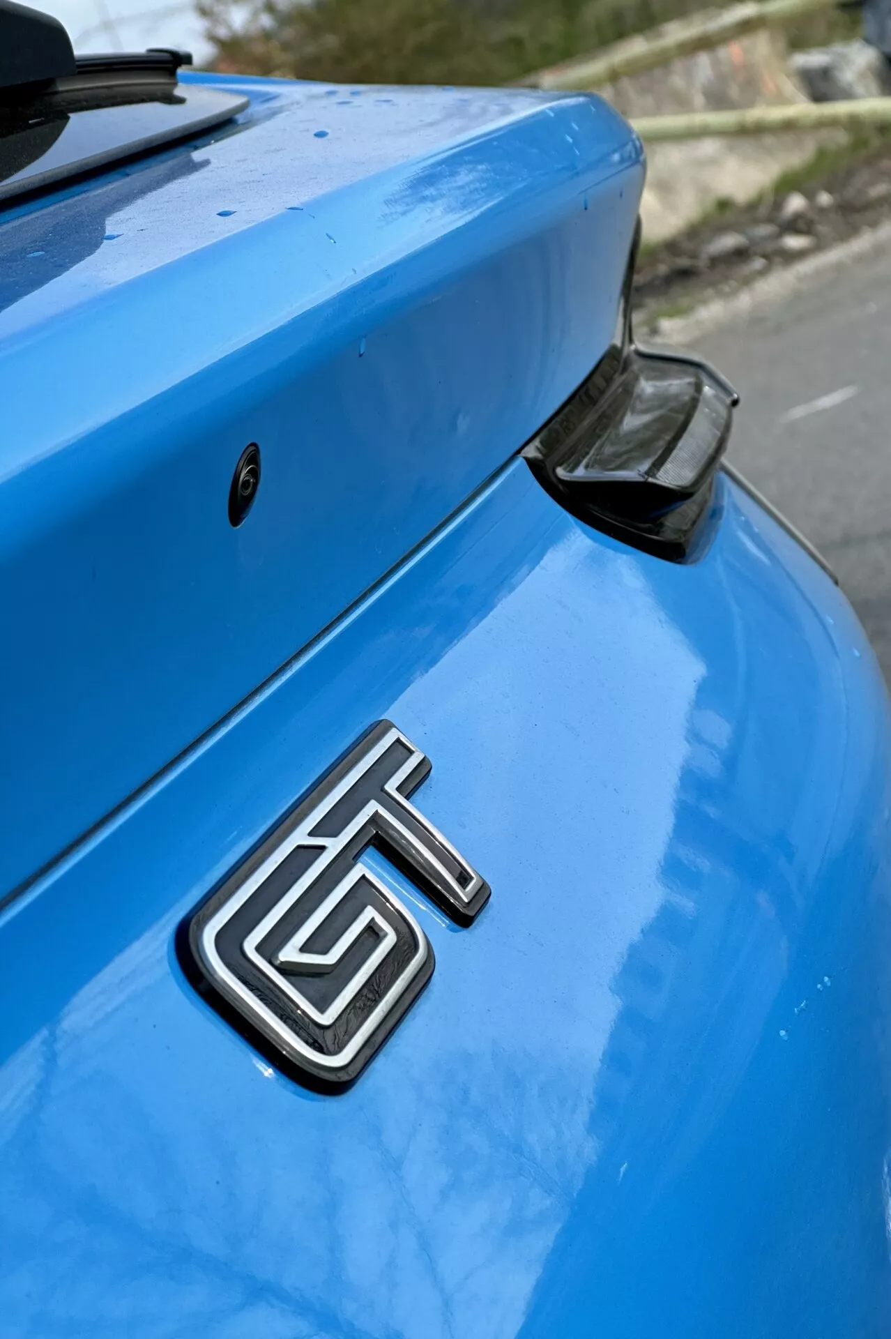 Close-up of a blue 2020 Ford Mustang Mach-E focusing on the rear section with detailed view of the metallic GT badge, curved trunk, and aerodynamic rear spoiler with a black end cap, against a softly blurred background.
