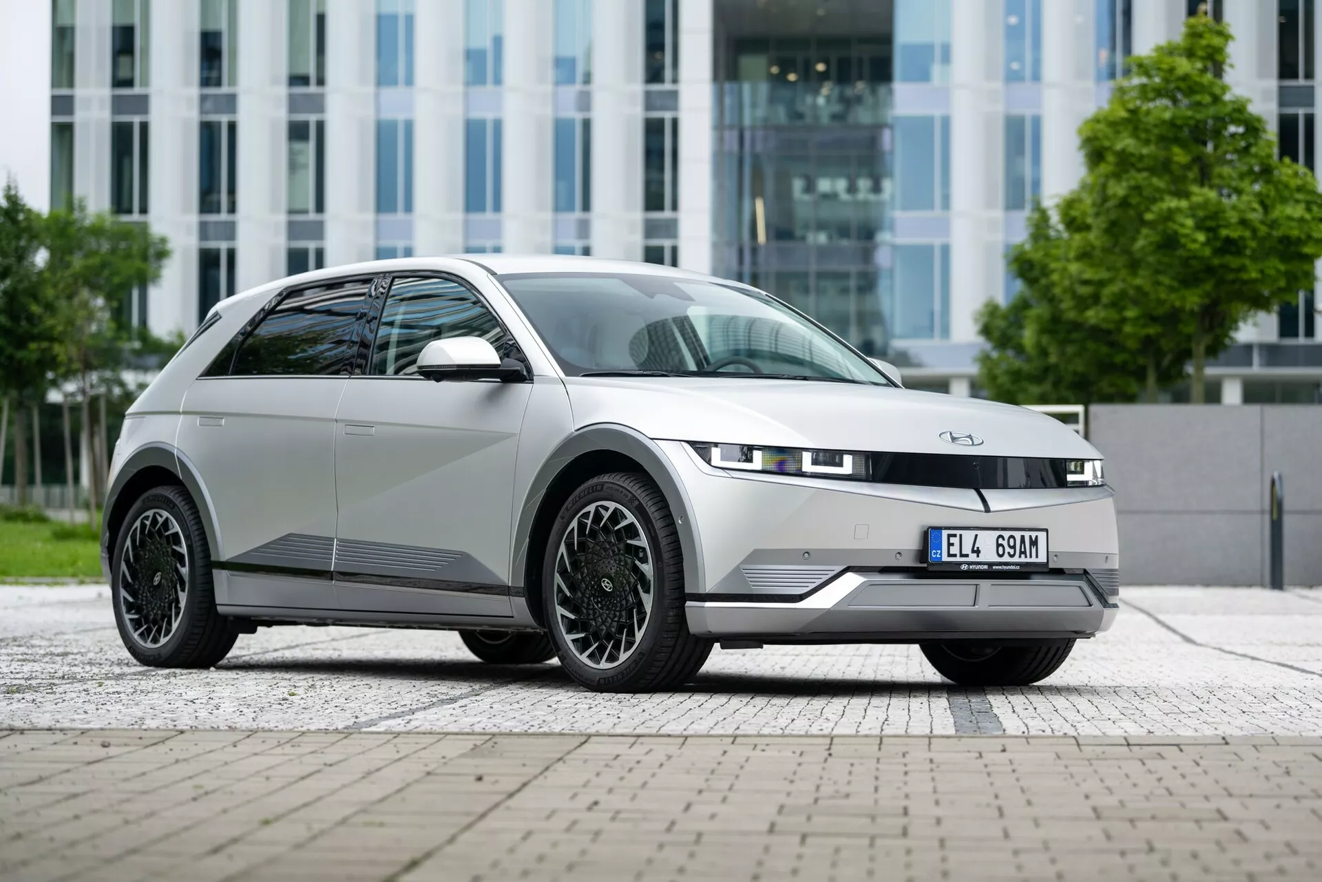 A silver-gray 2021 Hyundai Ioniq-5 2WD electric car captured from a low, frontal angle, emphasizing its stylish front right side. The vehicle is parked on a paved area with rectangular stone tiles in front of a modern building with a glass facade. The focal point highlights the sleek, minimalist grille and slim LED headlights. The hood slopes smoothly towards the front badge, complemented by a contrasting black trim on the bumper. The intricate black multi-spoke wheels and the character line running along the side contribute to the dynamic, modern, and eco-friendly design of the car.