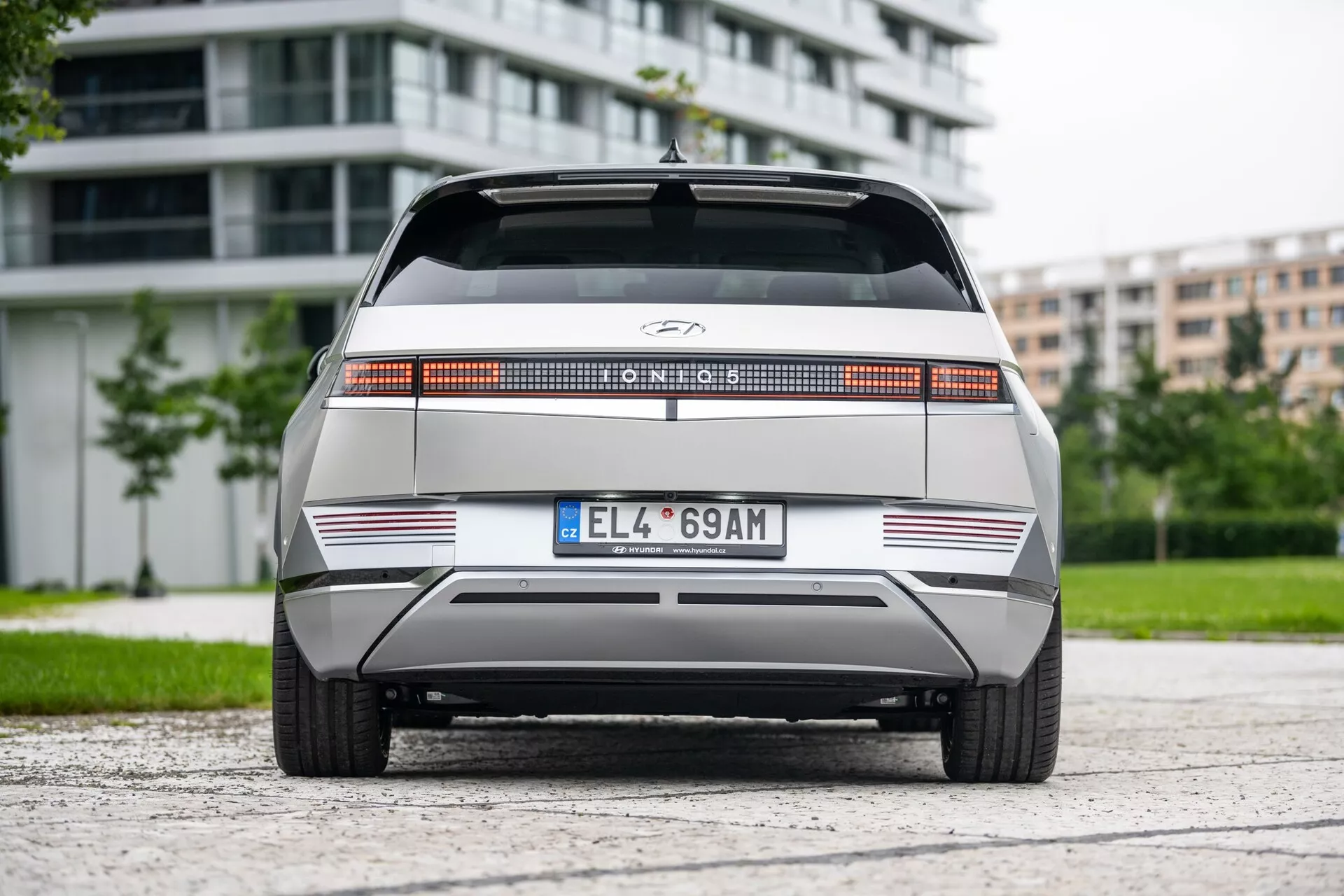 Rear view of the 2021 Hyundai Ioniq 5 2WD, highlighting a distinctive full-width rear light bar, centrally located manufacturer's logo, flat rear with sharp horizontal lines, minimalistic rear bumper with reflective strips, and European-style license plate. The vehicle is centered on an asphalt surface with foliage and modern buildings in the background, under overcast lighting.