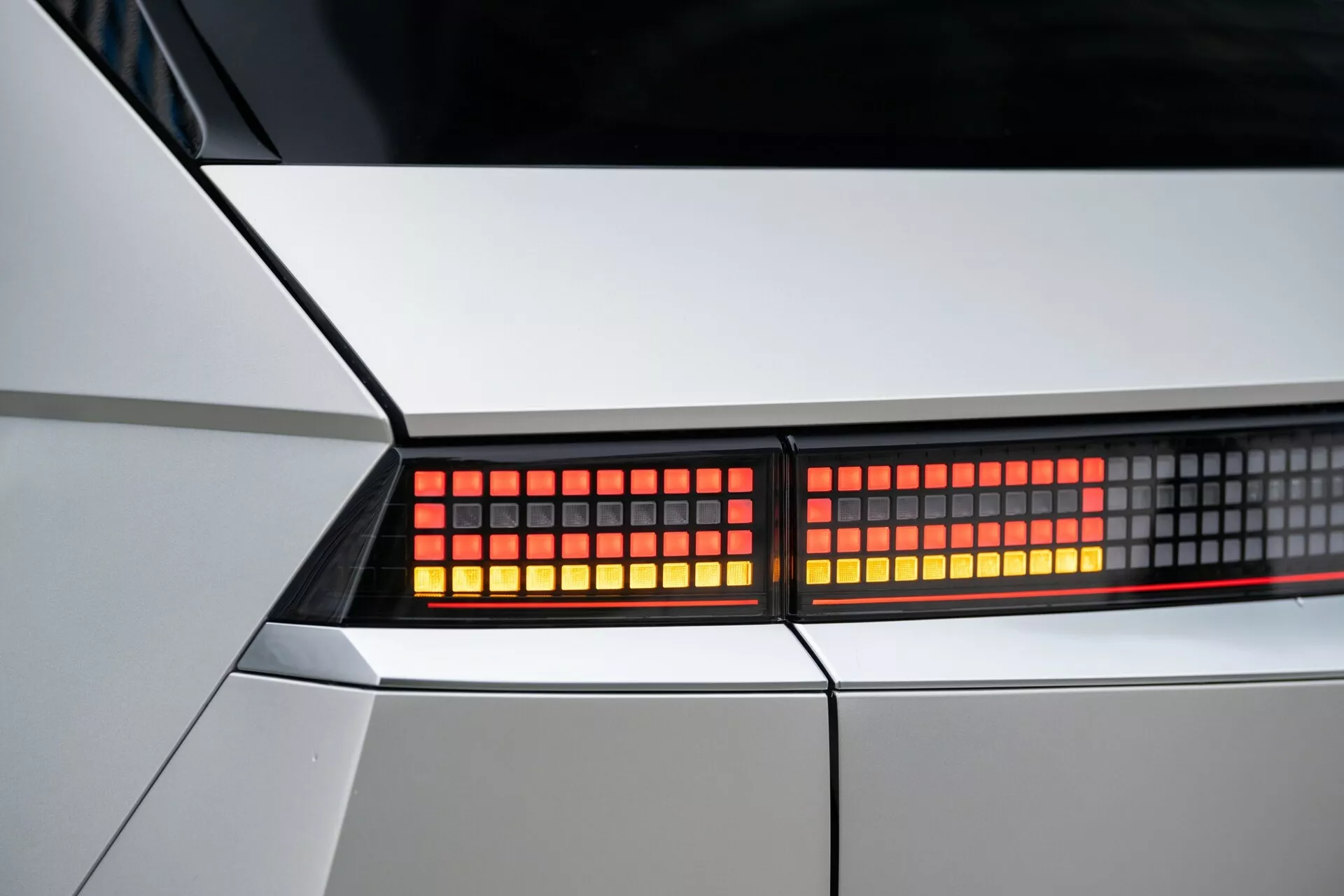 Close-up image of Hyundai Ioniq-5 2021 2WD rear taillight assembly. Focused shot on red, pixelated LED lights in a tight crop view, emphasizing the car's sharp, geometric bodywork in white or light silver. Detailed view of modern, futuristic design elements and lighting technology.