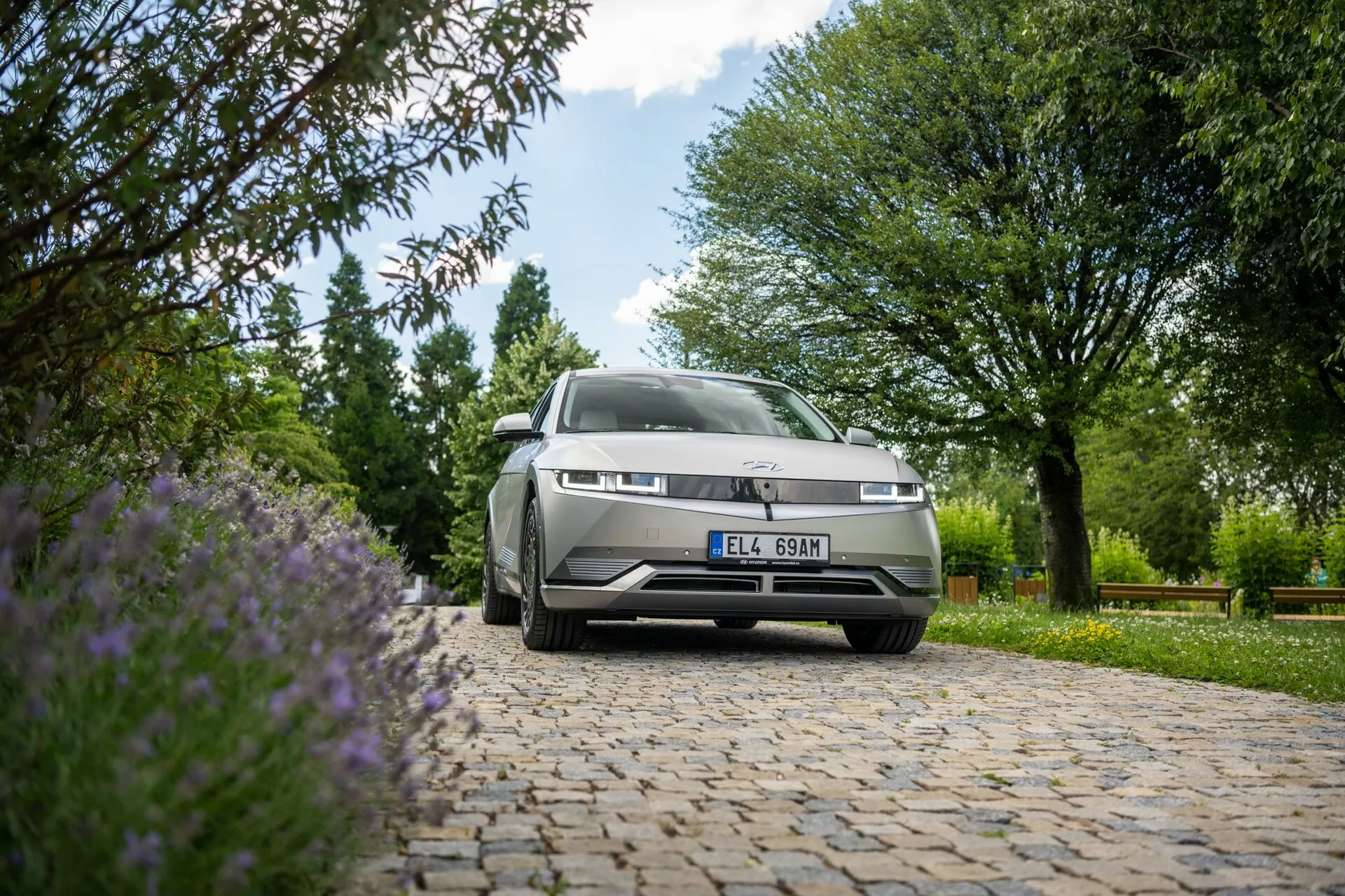 Low-angle shot of a Hyundai Ioniq 5 2WD 2021 parked outdoors on a cobblestone surface. The camera focuses on the front end of the car including the grille, left headlights, and part of the hood and windshield, set against a backdrop of green trees, bushes, and purple flowers, in ample natural daylight.