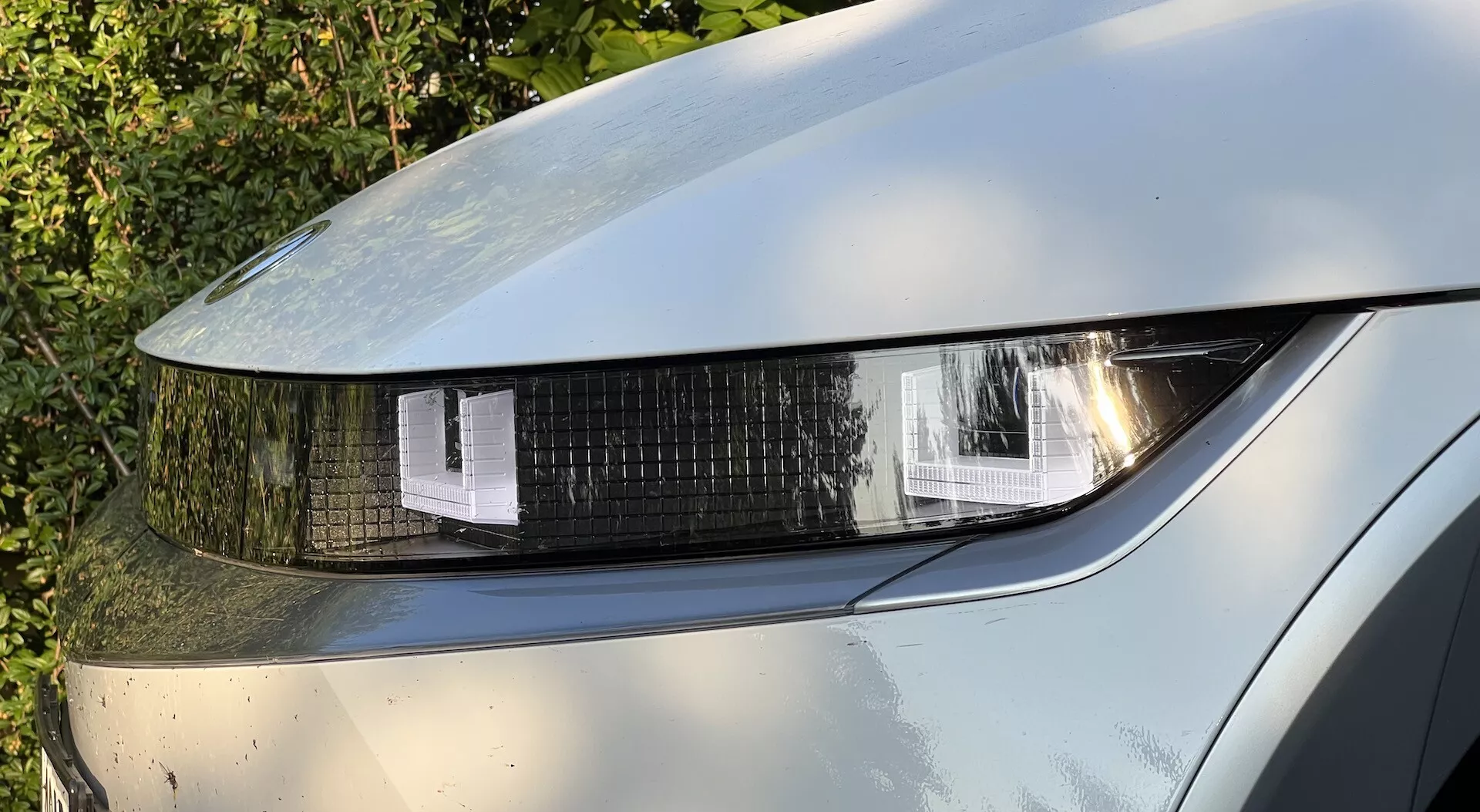 Close-up view of a 2021 Hyundai Ioniq-5's front left headlight, showcasing LED or HID lighting with geometric shaped segments, a shiny clear cover, and reflections on metallic paint with visible dirt, indicating usage.