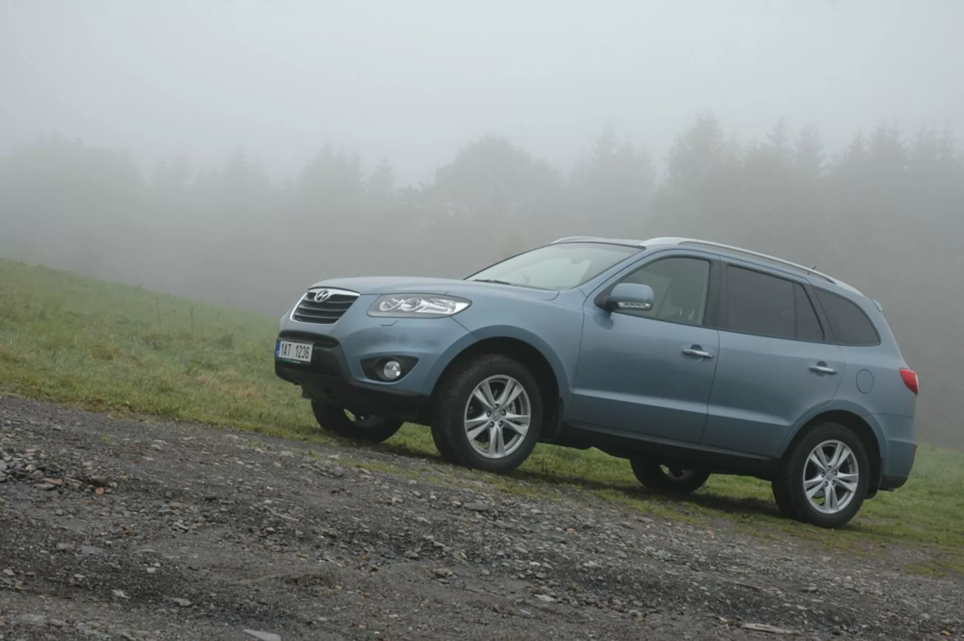 2006 Hyundai Santa Fe blue mid-size crossover SUV on an unpaved road with a grass patch, angled front-left view in foggy weather highlighting its robust design, off-road capabilities, and modern family vehicle features like front grille, angular headlights, fog lights, alloy wheels, roof rails, and integrated turn signals.