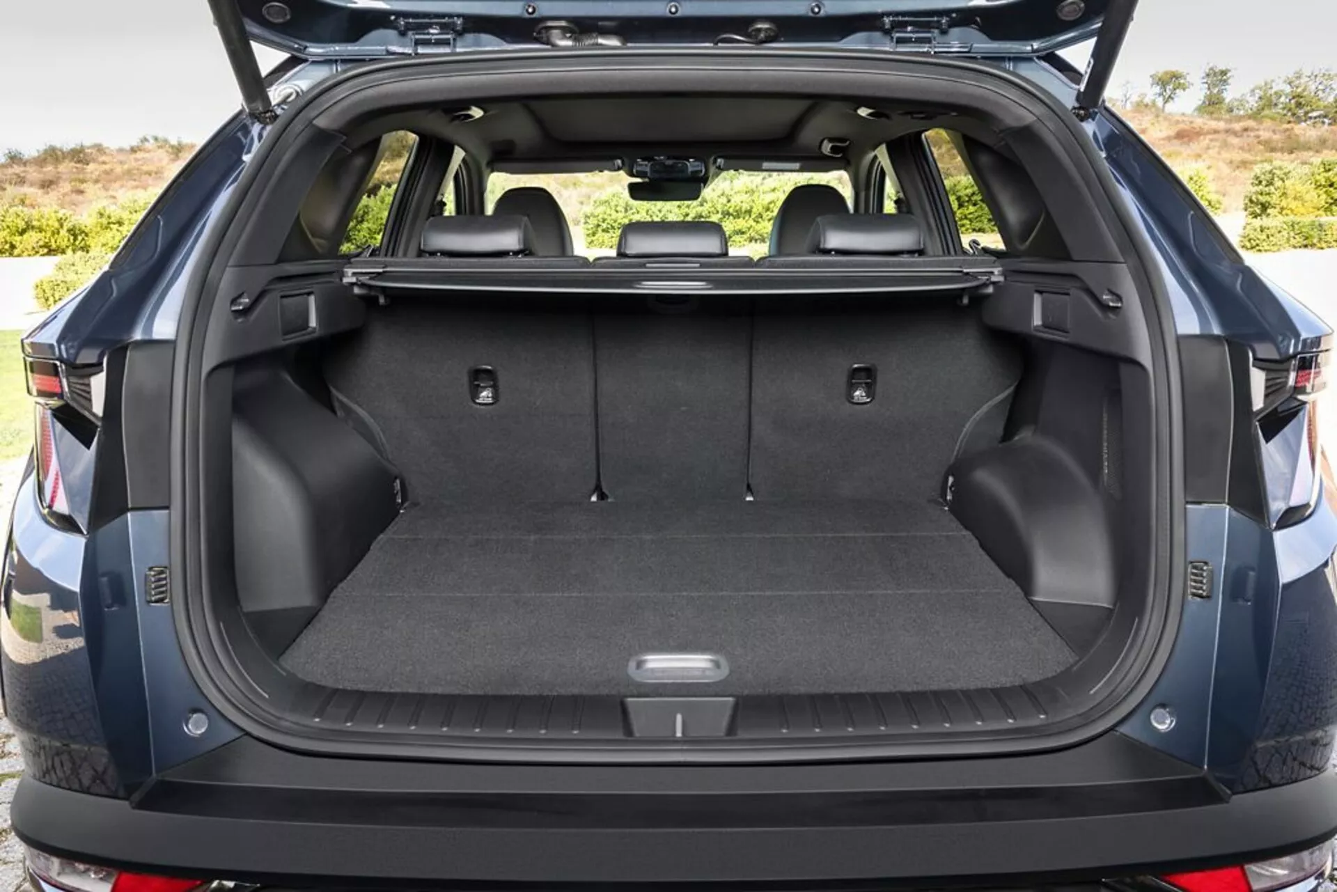 2020 Hyundai Tucson rear cargo area with trunk open, showcasing carpeted load floor, tethering points, storage compartments, 12V power outlet, foldable rear seats, retracted cargo cover, durable plastic sidewalls, and a glimpse of the rear bumper, in an outdoor setting.