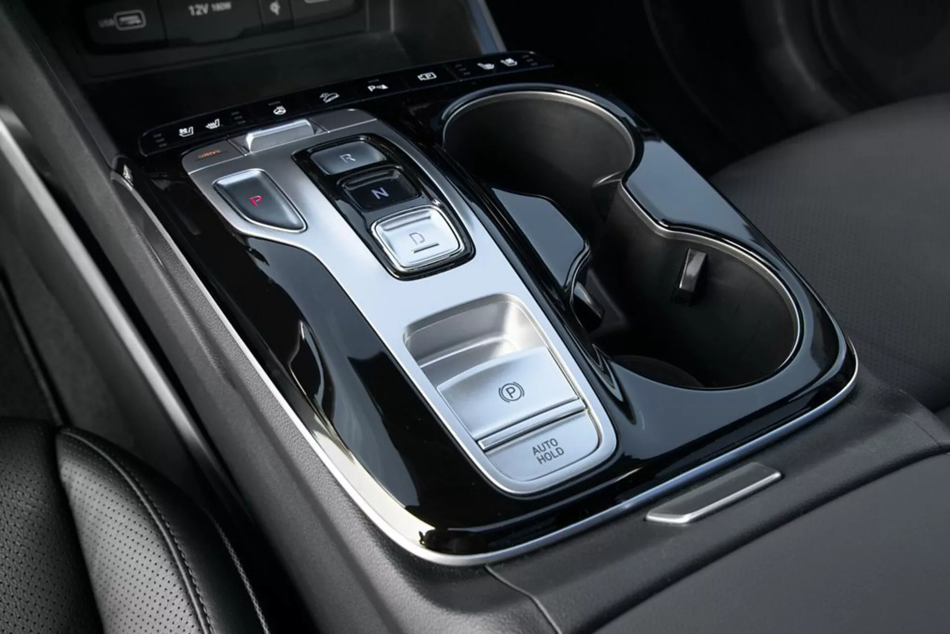 2020 Hyundai Tucson center console close-up showing gear shift lever, control buttons, Auto Start/Stop function, hazard light switch, and dual-cup holders with chrome accents, from a top-down view with slight incline.
