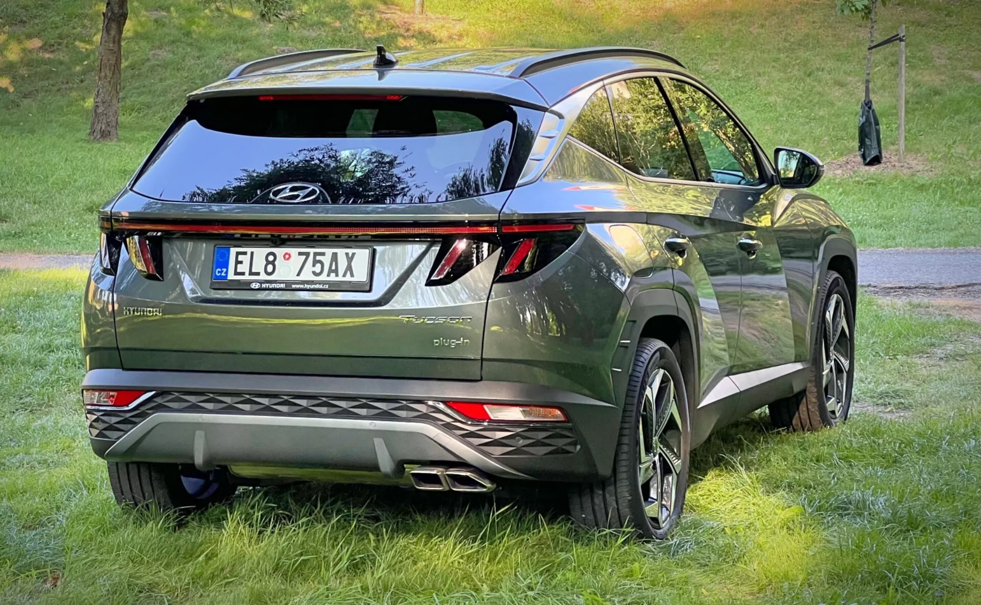Three-quarter rear view of a 2020 Hyundai Tucson, showcasing its LED tail lights, aerodynamic design with a roof spoiler and shark-fin antenna, premium dark metallic green finish, and equipped with large alloy wheels and parking sensors, in a crossover urban lifestyle setting