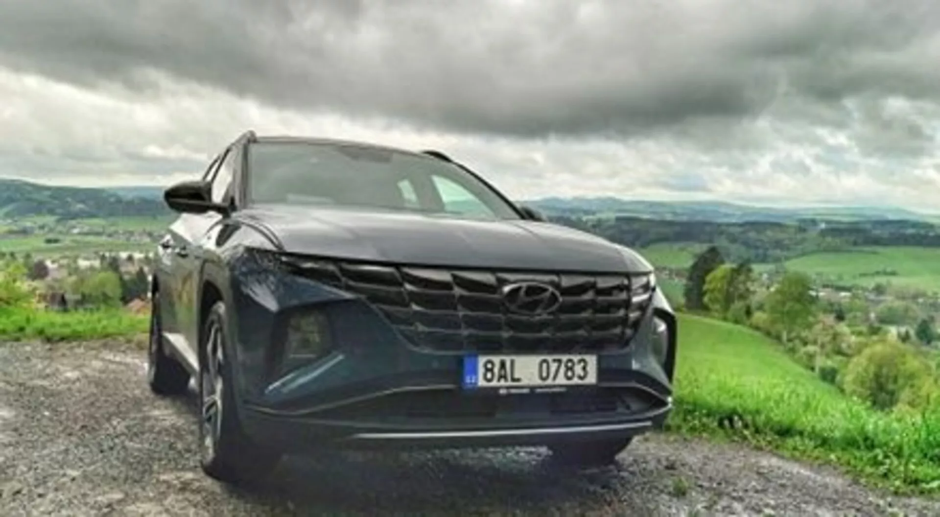 2020 Hyundai Tucson SUV parked on gravel road, showcasing its bold grille, sleek LED headlights, and metallic paint in a dynamic low front angle view, with a lush hilly landscape under a cloudy sky in the background