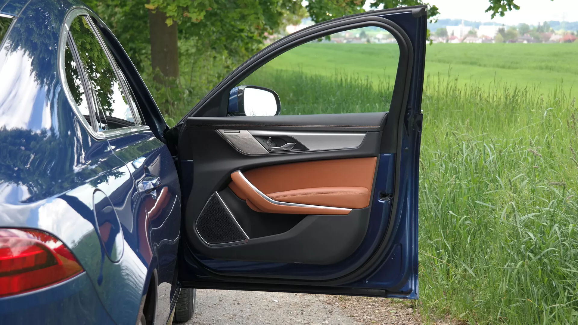 Close-up lateral view of a 2021 Jaguar XF with the driver's side door open, revealing a luxurious interior with a combination of black and warm tan leather. The side mirror, integrated with a turn signal, matches the car's sleek blue exterior. Parked on a paved surface near a grassy field, the car's partially visible fender and quarter panel highlight its modern design and advanced technological features.