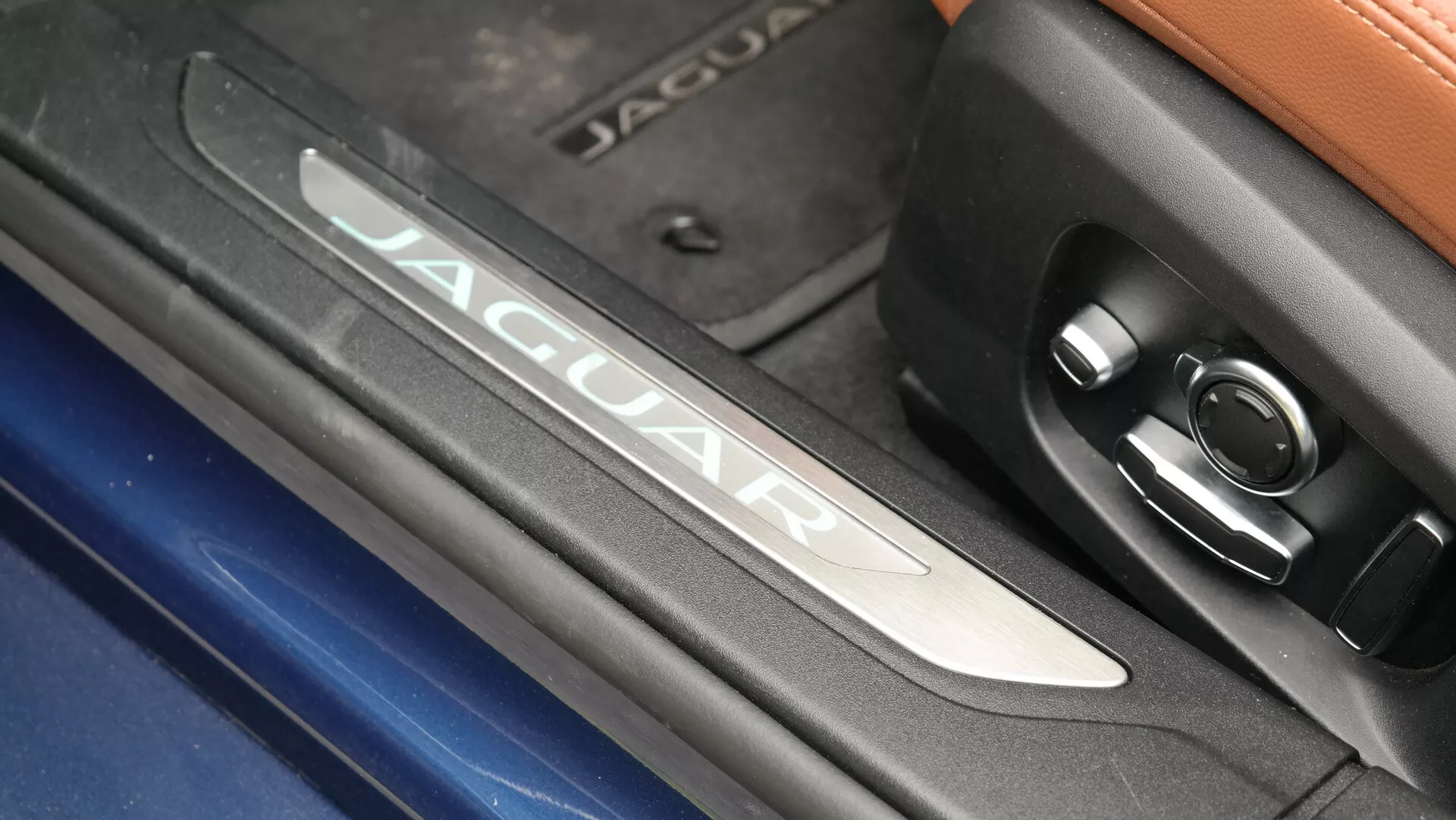 Close-up detail of the Jaguar XF 2021 interior, highlighting the illuminated 'JAGUAR' branding on the brushed metal door sill plate, adjacent to circular patterned design and a control button. Contrasting textures include chrome finish, blue exterior glimpse, and leather upper section in a luxurious, high-tech vehicle environment.
