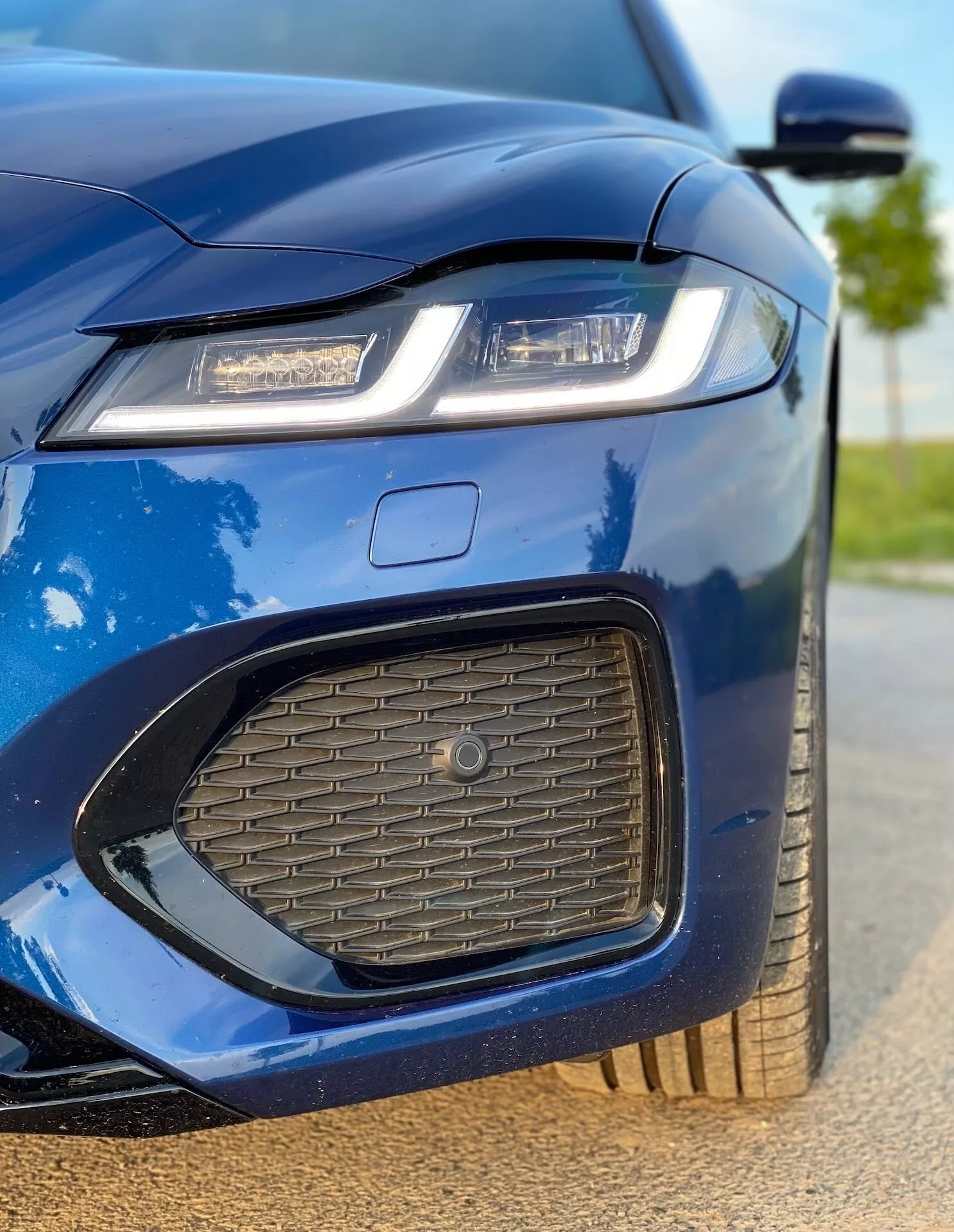 Close-up image of the front left section of the 2021 Jaguar XF in metallic blue. The shot captures the left headlight with modern LED or laser lighting technology, a part of the hood, the front bumper with a honeycomb mesh air intake, a section of the grille with a large emblem, and a bit of the left wing mirror. The image highlights the car's sharp, aggressive design, sporty aesthetics, polished finish, aerodynamically designed side mirror with integrated turn signals, and partially visible alloy wheel, showcasing the car's luxury and technological features.