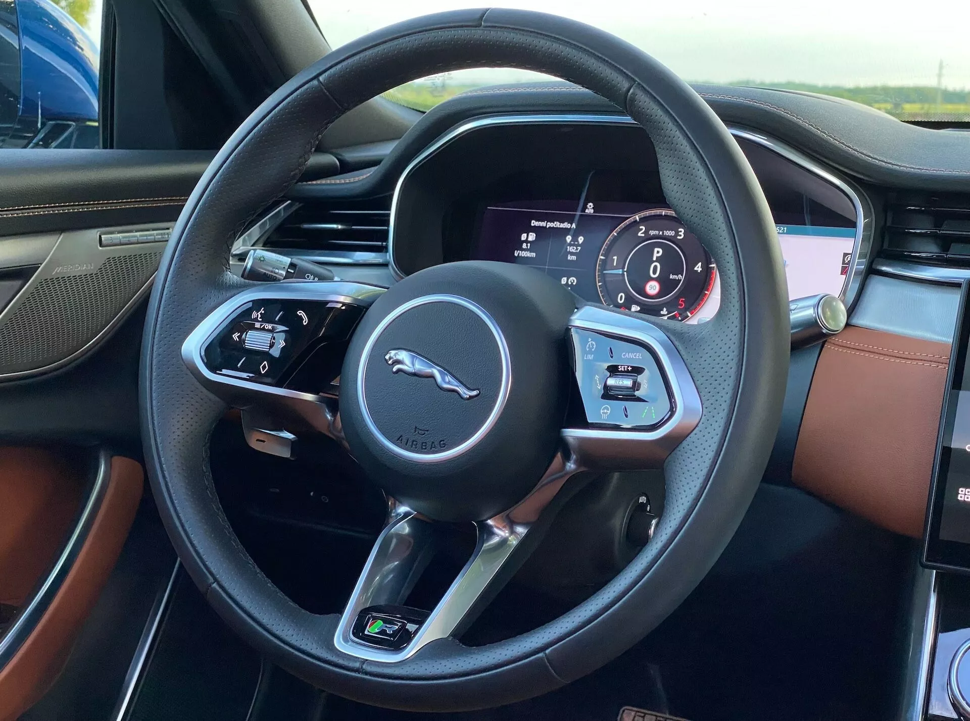 Interior view of the Jaguar XF 2021 from the driver’s seat, showcasing the three-spoke black leather-wrapped steering wheel with visible stitching, featuring the iconic leaping feline logo. The left spoke contains controls for audio and voice commands, while the right spoke includes cruise control and driver information system buttons. The instrument cluster behind the wheel displays a mix of digital and analog gauges, including a speedometer and tachometer. The luxurious interior highlights two-tone leather upholstery in rich brown and black.
