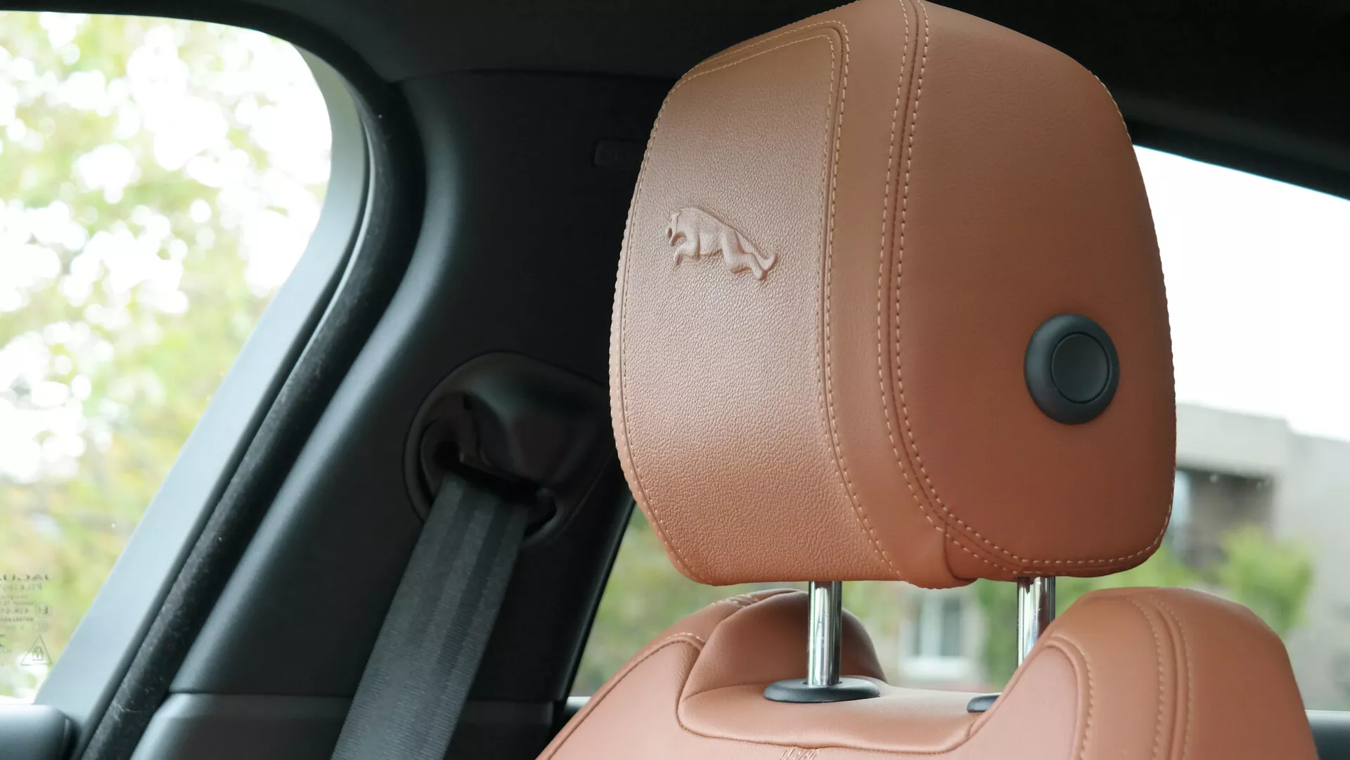 Interior view of the 2021 Jaguar XF focusing on the driver's side headrest, captured from a backseat perspective. The headrest, upholstered in high-quality tan or light brown leather with visible stitching, features an embossed logo, an ergonomic seatbelt guide, and a manual adjustment mechanism. The background includes a blurred B-pillar and window, highlighting the car's luxury and ergonomic design.