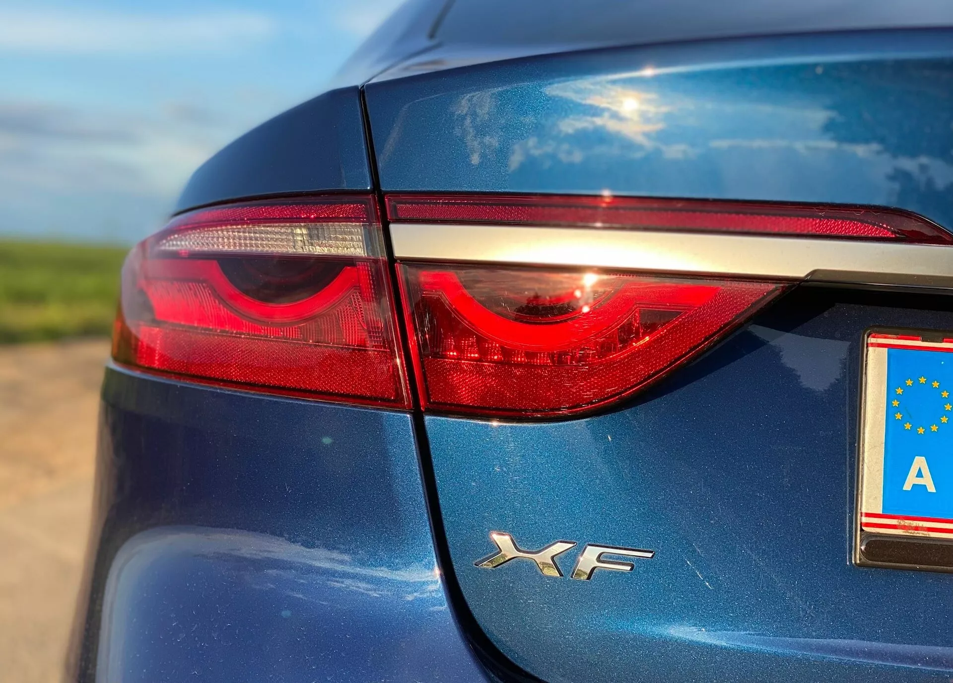 Close-up image showcasing the rear left section of a Jaguar XF 2021. The photograph highlights the sleek, dark blue body, glossy finish, and left taillight, featuring an LED light strip. The EU license plate displays an 'A' for Austria, with reflections indicating natural daylight.