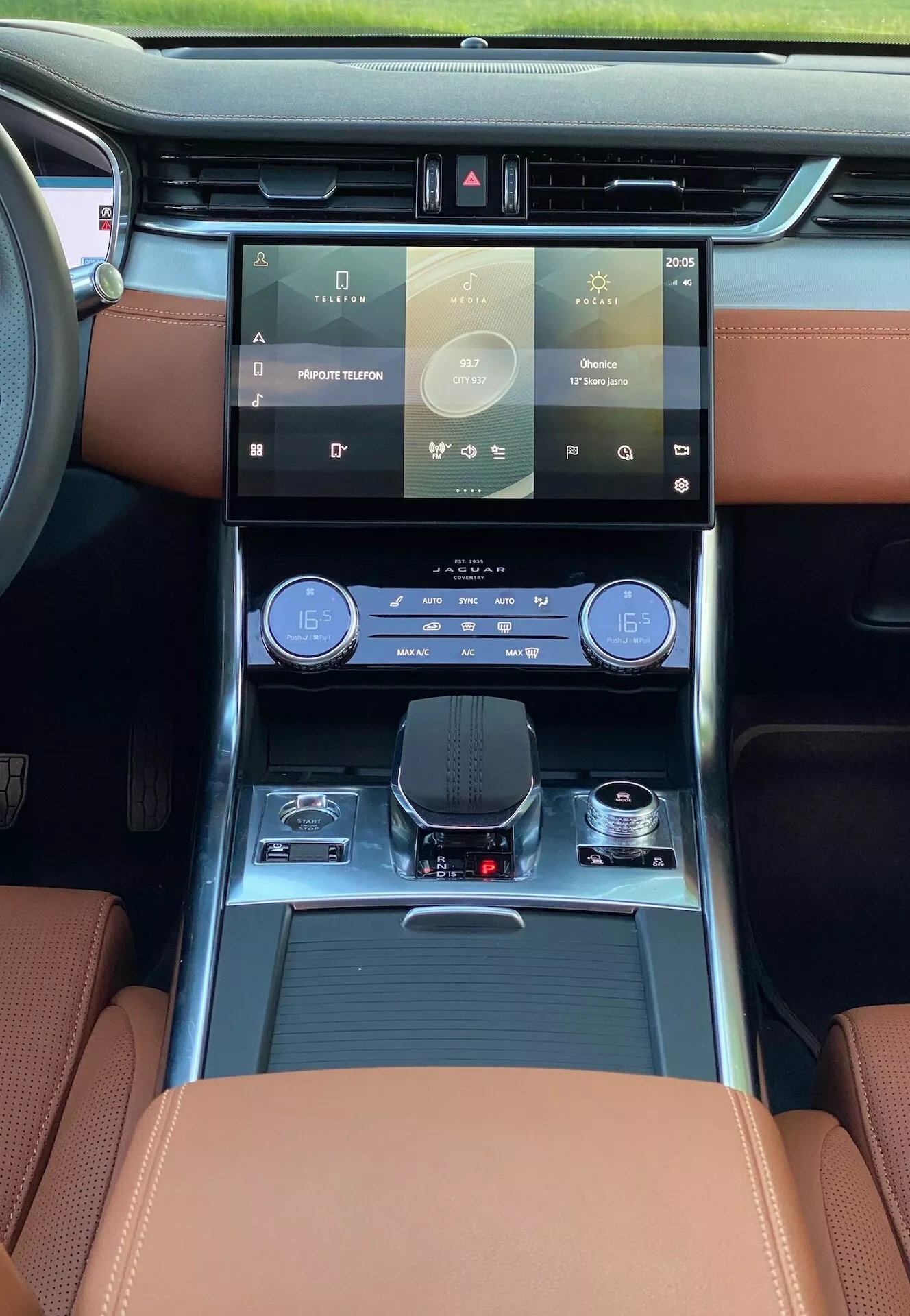 Interior of the 2021 Jaguar XF, showcasing a driver-centric perspective from the front seat with focus on the dashboard and cutting-edge features. Highlights include a large, user-friendly touchscreen infotainment system, sleek dual-zone climate control panel, electronic shift-by-wire gear selector, luxury leather seating with contrasting stitching, and ambient lighting. Practical elements like cup holders and storage compartments are also visible. The natural daylight flooding the cabin suggests a panoramic sunroof or large windshield, enhancing the overall luxurious ambiance.