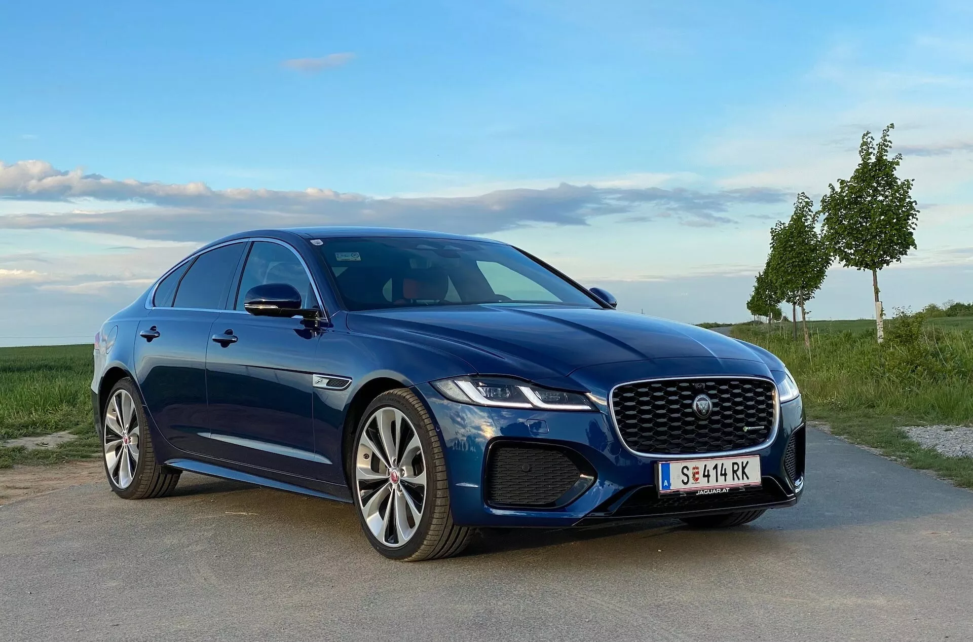 Blue Jaguar XF 2021 sedan parked on paved surface with grassy landscape and clear sky, showcasing distinctive grille, LED headlights, stylish front bumper with air intakes, and multi-spoke alloy wheels. The car’s sporty profile is enhanced by sleek lines and scooped side skirts, viewed from a low-angle three-quarter front perspective.