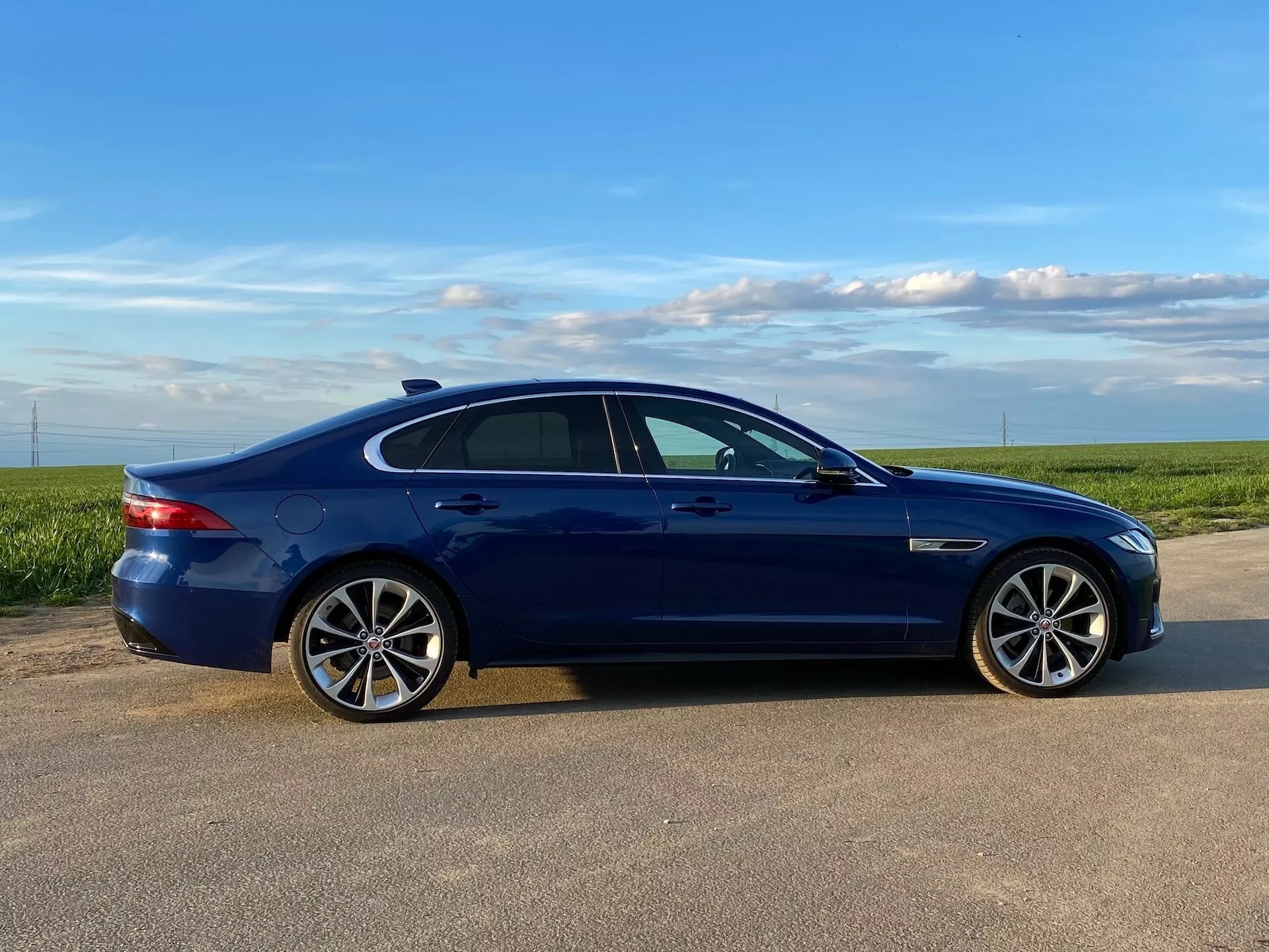 A side profile view of a blue 2021 Jaguar XF sedan captured in daytime lighting, showcasing its design features such as multi-spoke alloy wheels, low-profile tires, tinted windows, body-colored side mirrors with integrated turn signals, flush body-colored door handles, and chrome or brushed metal trim along the window line. The car is positioned on a paved surface with an open field and clear sky in the background, suggesting a rural or semi-rural setting.