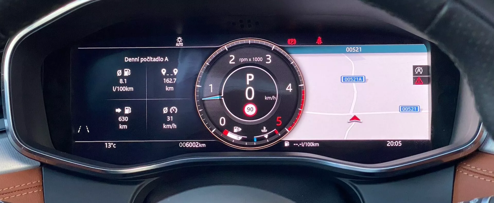 Close-up view from the driver's seat of a 2021 Jaguar XF's digital instrument cluster, featuring fuel economy metrics, an analog-style tachometer with RPM and digital speedometer, navigational information, odometer, external temperature, and time displayed. The cluster is framed within a dashboard with leatherette stitching, highlighting the luxury and technology aspects of the vehicle's interior.