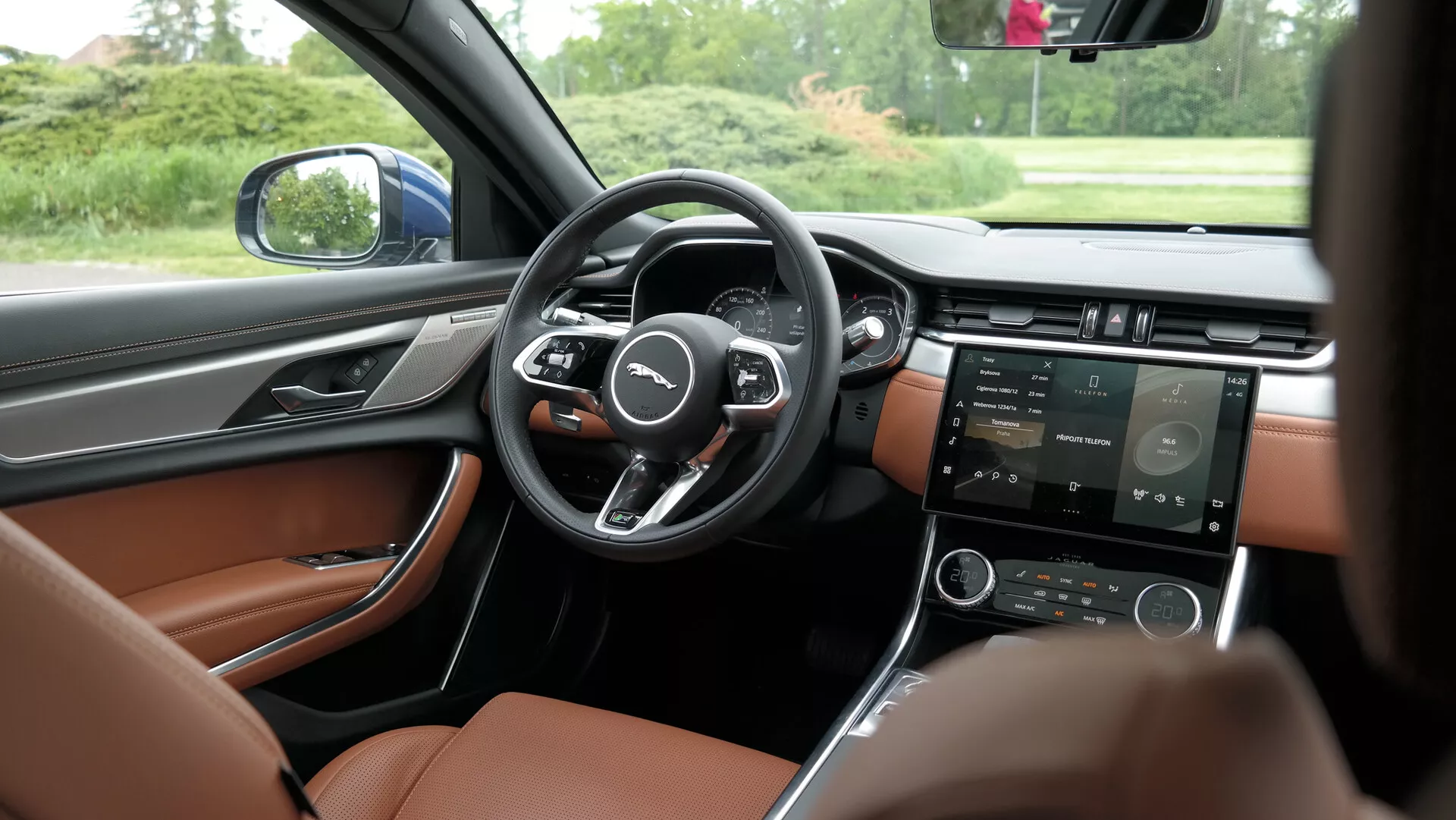 Interior view of a modern car from the perspective of the back seat, slightly offset to the driver's side, showcasing the front dashboard, steering wheel, center console, and driver's door. The steering wheel with a three-spoke design has multifunction buttons on each side. The digital instrument cluster behind it indicates configurable displays for driver information. The center console features a touchscreen for navigation, media, and vehicle settings, along with physical buttons likely controlling the climate and additional settings. The interior contrasts dark dashboard with luxurious lighter brown leather seats. The driver's door includes controls for windows, side mirrors, and seat adjustments, indicating an advanced technological package. The vehicle is stationary, surrounded by trees as seen through the side window, emphasizing the clarity of the interior details.