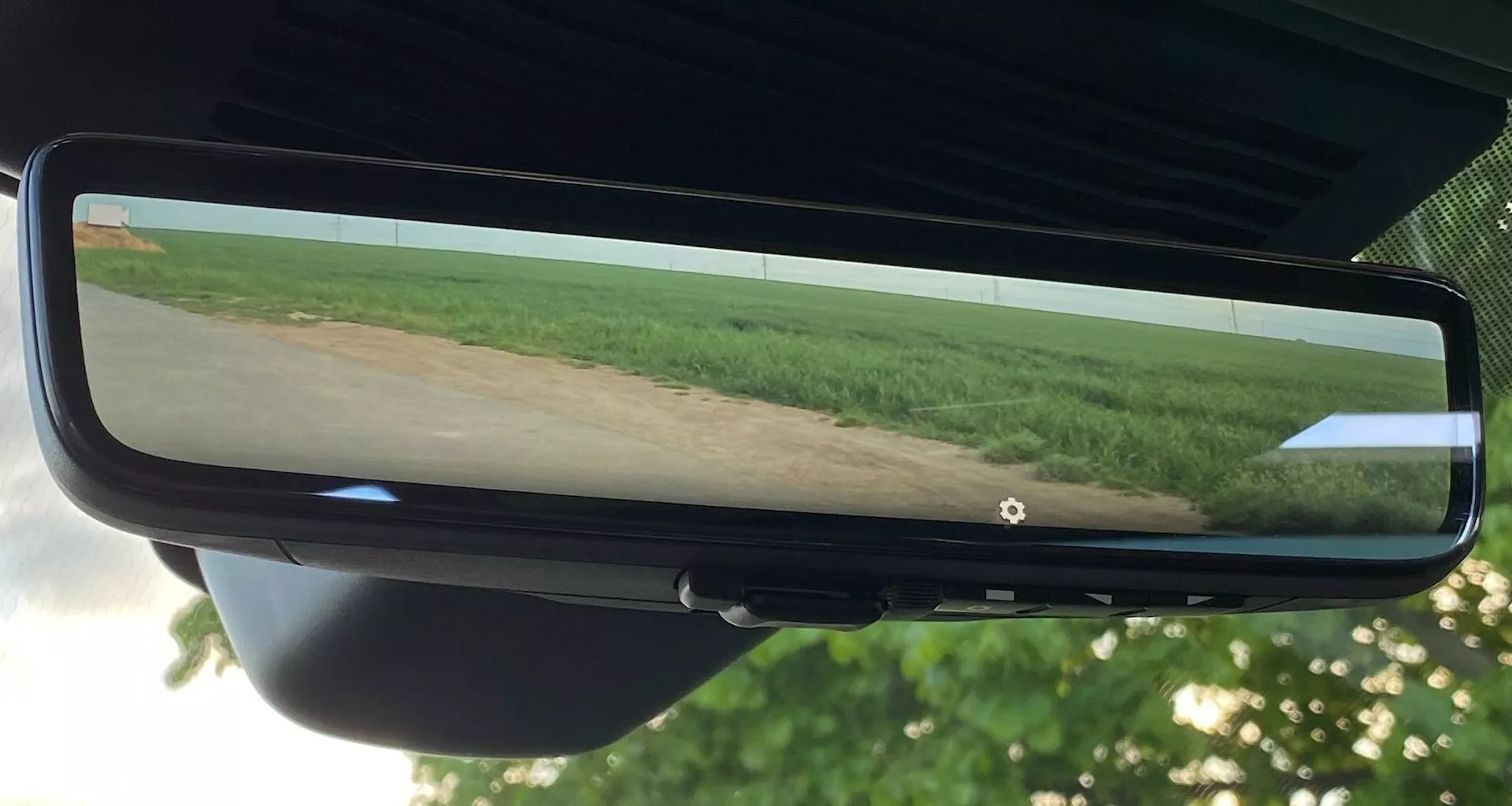 Rear-view mirror of the 2021 Jaguar XF reflecting a wide-angle view of a field with vegetation and clear sky, centered in the photo. The mirror is attached to the windshield mount and surrounded by a black bezel, with sections of the vehicle's headliner and rear interior visible.