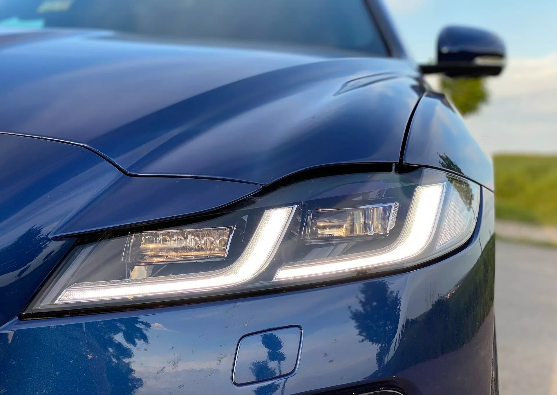 Close-up view of the 2021 Jaguar XF's front-left section with a low, off-center angle highlighting the modern left headlight with distinctive LED daytime running lights. The deep blue glossy paint and sculpted hood with aerodynamic crease lines are prominently displayed. The side mirror is partially visible, adding to the sleek aesthetic. The shallow depth of field emphasizes the car's design and advanced lighting system.