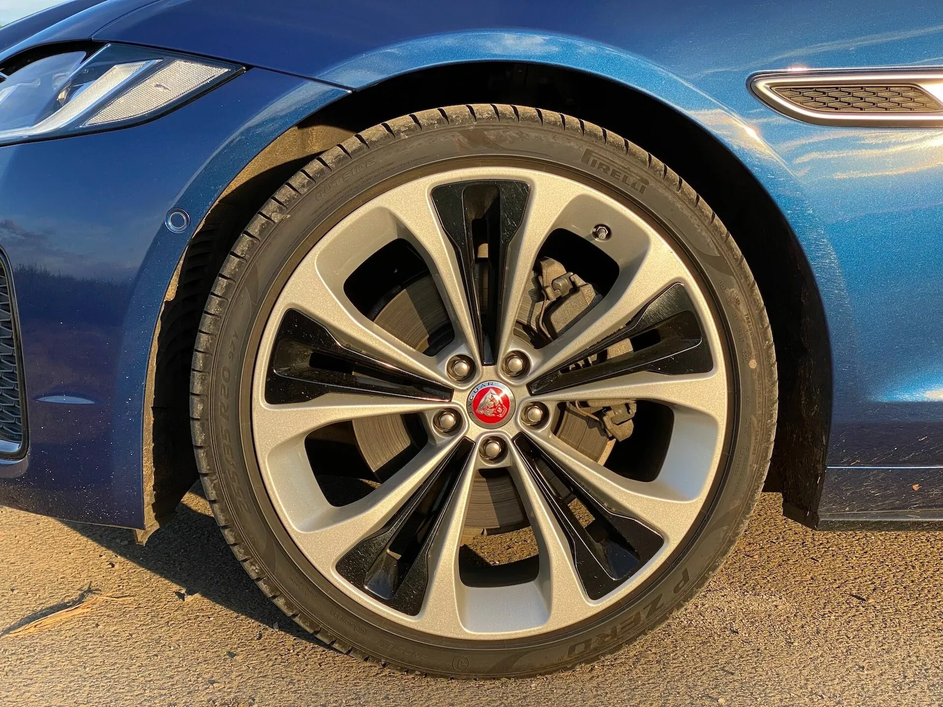 Close-up view of a 2021 Jaguar XF front passenger-side wheel from a low angle. The multi-spoke alloy wheel features a dynamic design with ten pairs of thin and thick spokes. The center cap displays a red brand emblem. The on-road tire has a visible tread pattern, and behind the spokes, the brake caliper and ventilated disc rotor can be seen. The blue car body with a glossy finish part and curved fender are visible. The image highlights the car's luxury and style features, suitable for discussions on car parts, design details, and brand identification.