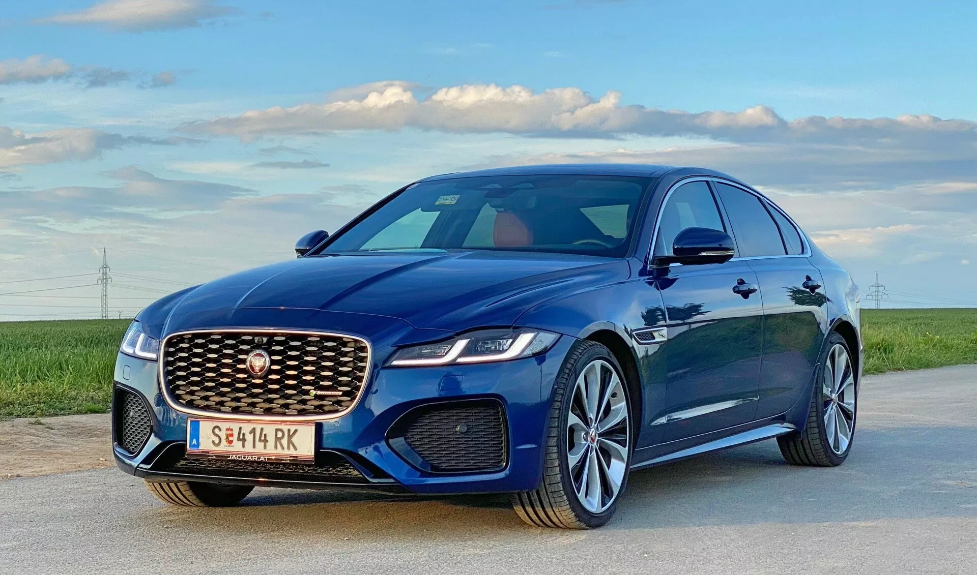 The image shows a modern luxury sedan, a 2021 Jaguar XF, parked on an open road beneath a clear sky with fields and distant power lines on the horizon. Captured from a low-angle, three-quarter front view emphasizing the driver's side, it highlights the prominent mesh-patterned front grille with central emblem, sleek LED headlights, sculpted hood lines, aerodynamic side mirrors, and multi-spoke alloy wheels with metallic paint. Features inferred include advanced LED headlights, active aerodynamics, alloy wheels with performance tires, and potential advanced driver assistance systems.