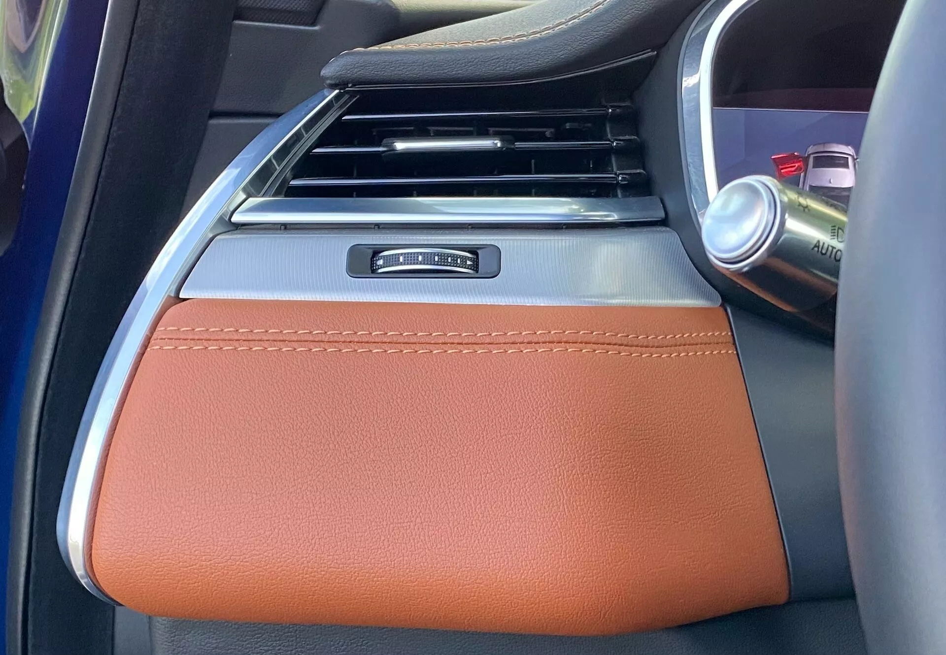 Interior detail of the 2021 Jaguar XF, focusing on the center armrest area. The image is taken from the rear seat looking forward, showing a brown leather armrest with visible stitching, suggesting a luxury trim level. The upper part of the armrest features a metallic trim piece, possibly decorative or functional. Behind the armrest, the lower part of the center console with vertical air vents, black with silver accents, can be seen. To the right, a cup holder with a can indicates additional storage. The partially visible silver-colored part likely indicates control for heating, cooling, or other comfort features, highlighting the modern, premium interior design.