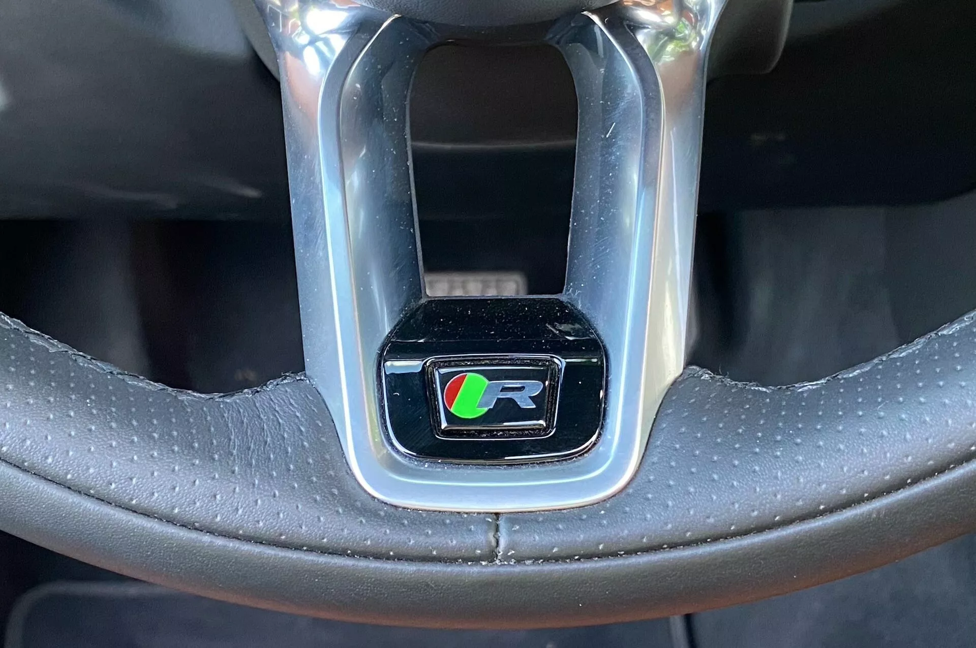 Close-up shot of a car's steering wheel section highlighting the ‘R’ performance line emblem encased in a shiny chrome rectangle. The emblem, with a black background, features a stylized ‘R’ with colored stripes in green, blue, red, and possibly yellow. The steering wheel is made of black leather, partly perforated for better grip and luxury feel, with visible stitching. The image has a front-facing, slightly upward angle, focusing solely on the emblem, excluding the rest of the car's interior.